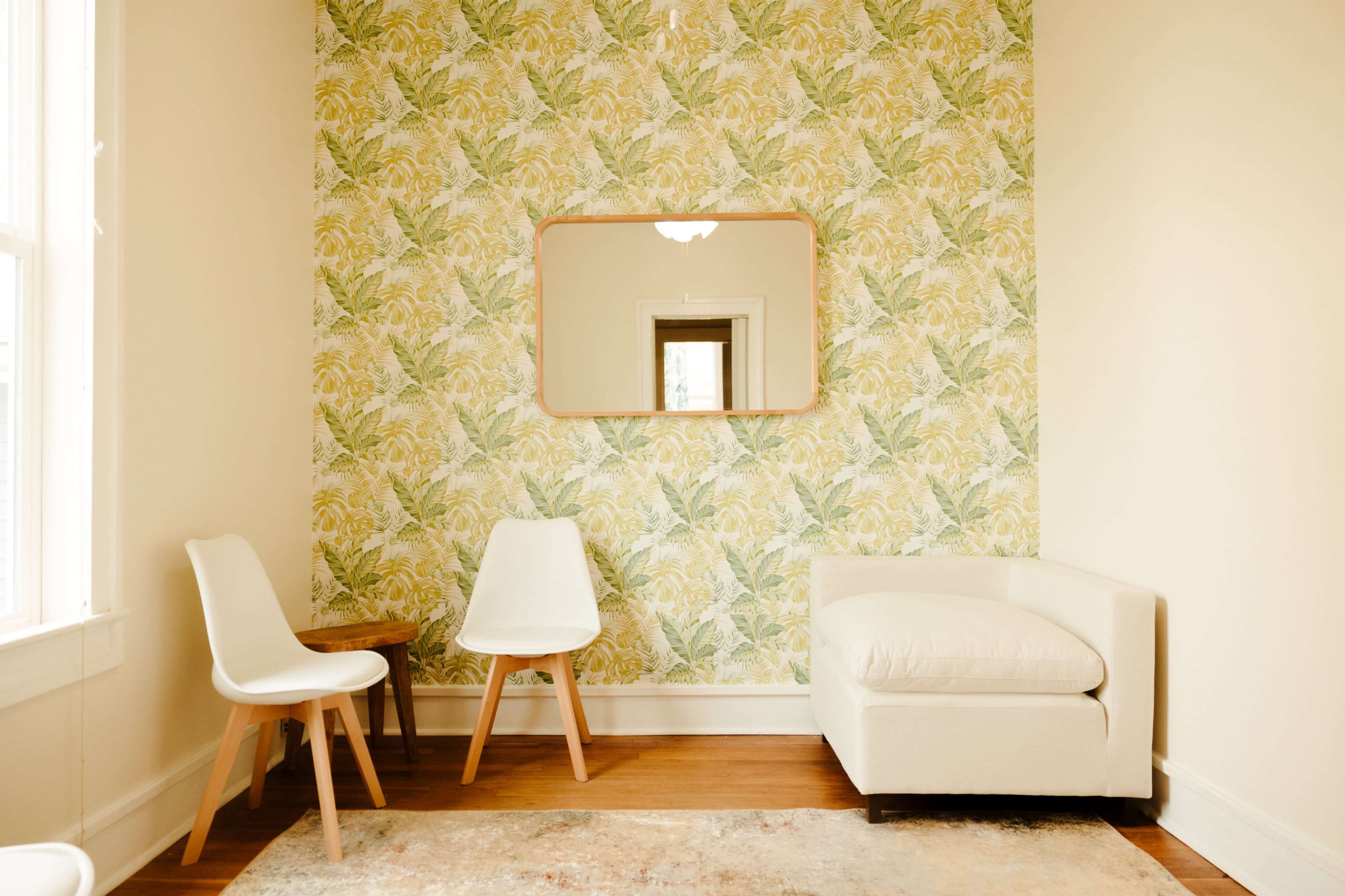 A small room features a patterned yellow and green wallpaper, a round mirror, a white armchair, two white chairs, and a small wooden table.