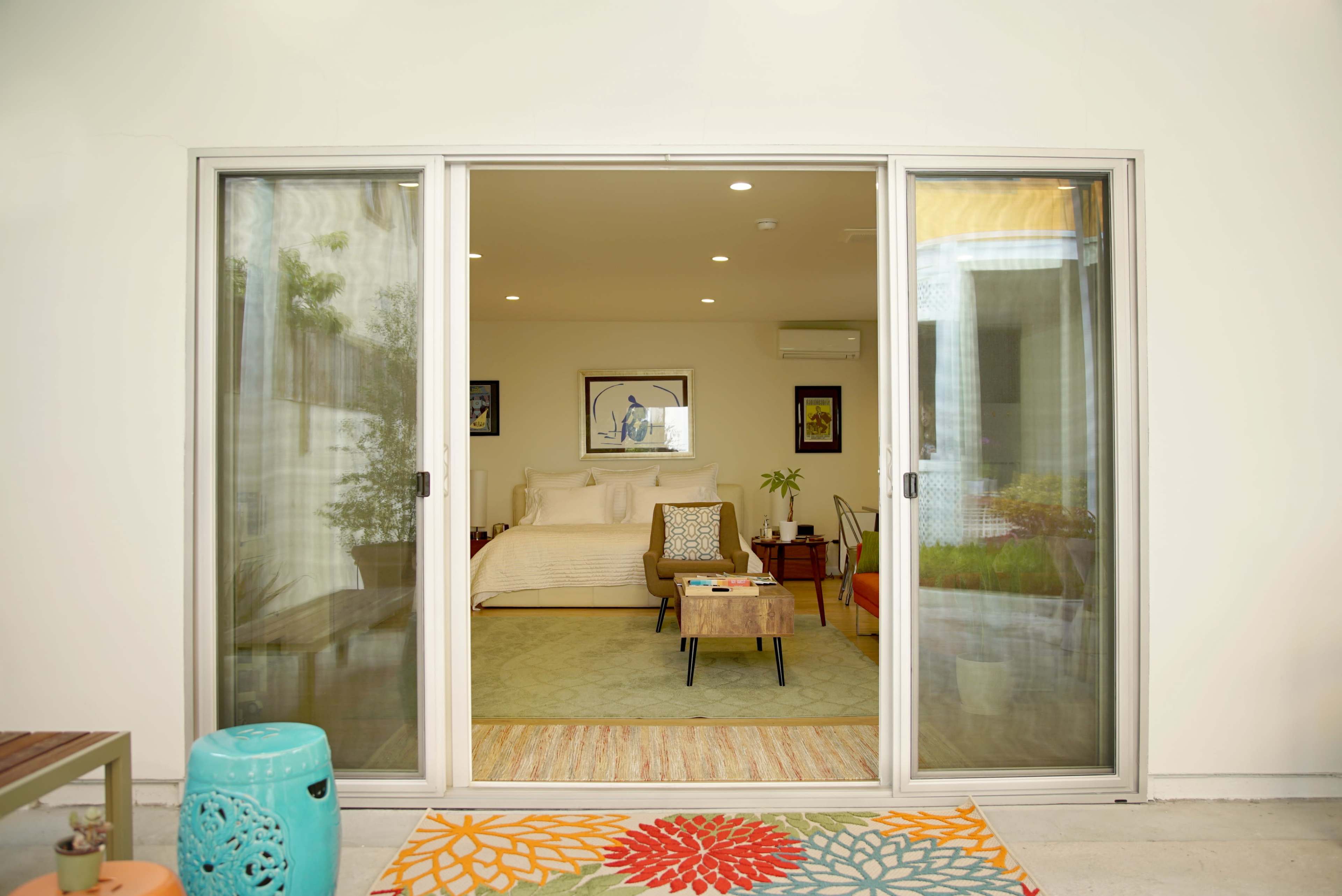 A view through sliding glass doors into a bright bedroom featuring a bed, seating area, and decorative plants.