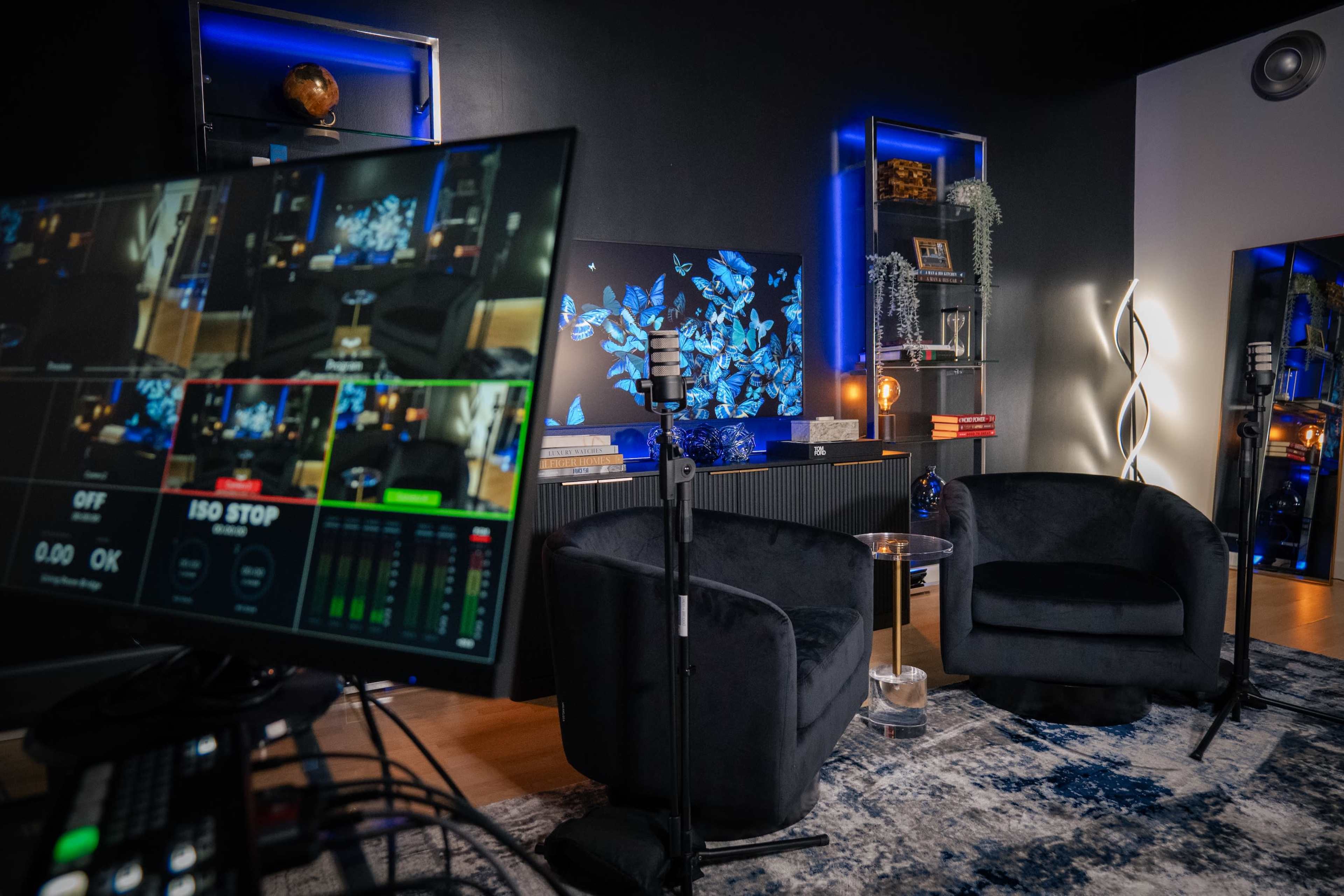 The image shows a modern recording studio setup with two black chairs, multiple microphones, and a large screen displaying colorful visuals in a dimly lit environment.