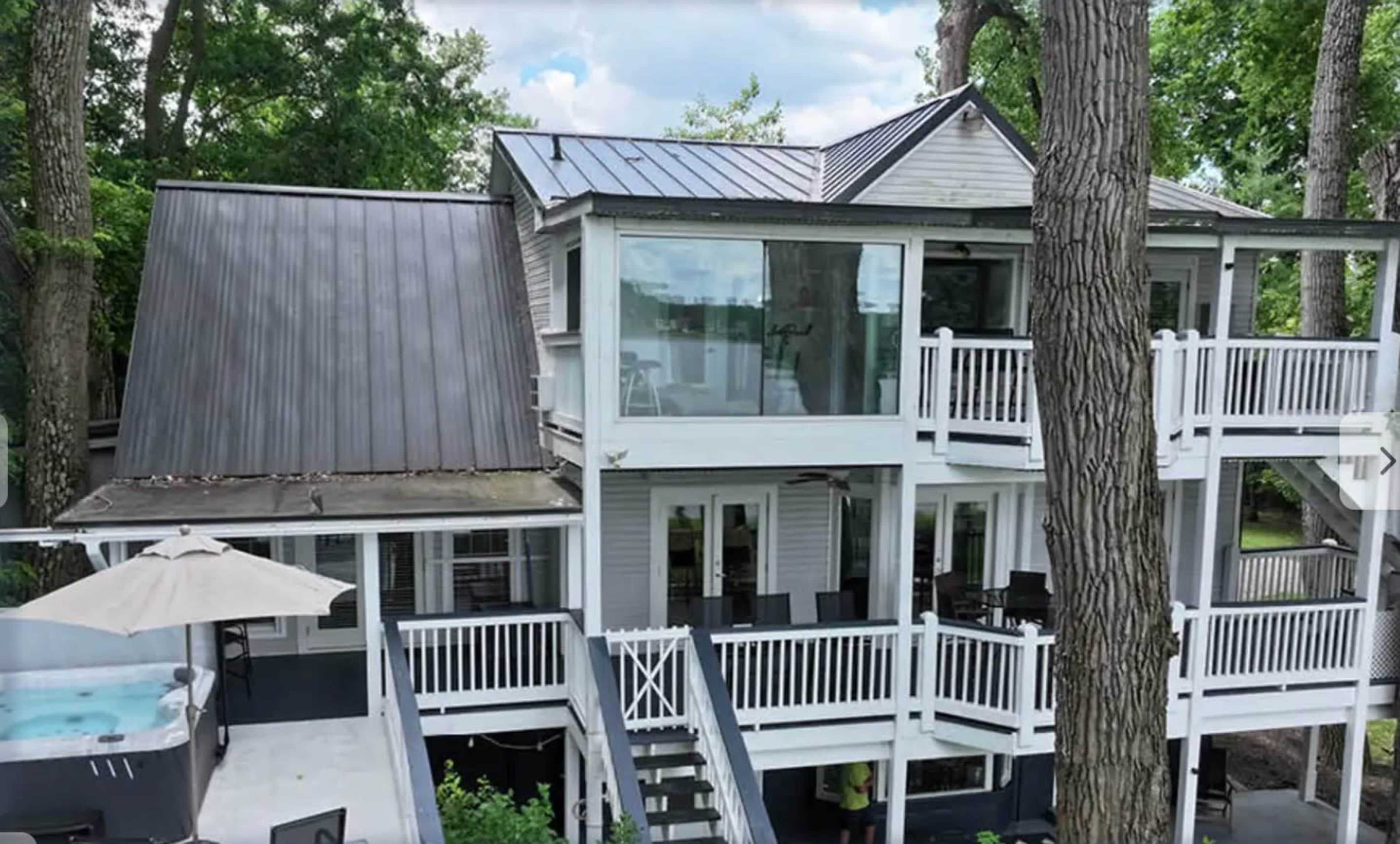 The image shows a multi-level modern house with a combination of metal and shingled roofs, surrounded by trees and featuring a balcony and outdoor seating area.
