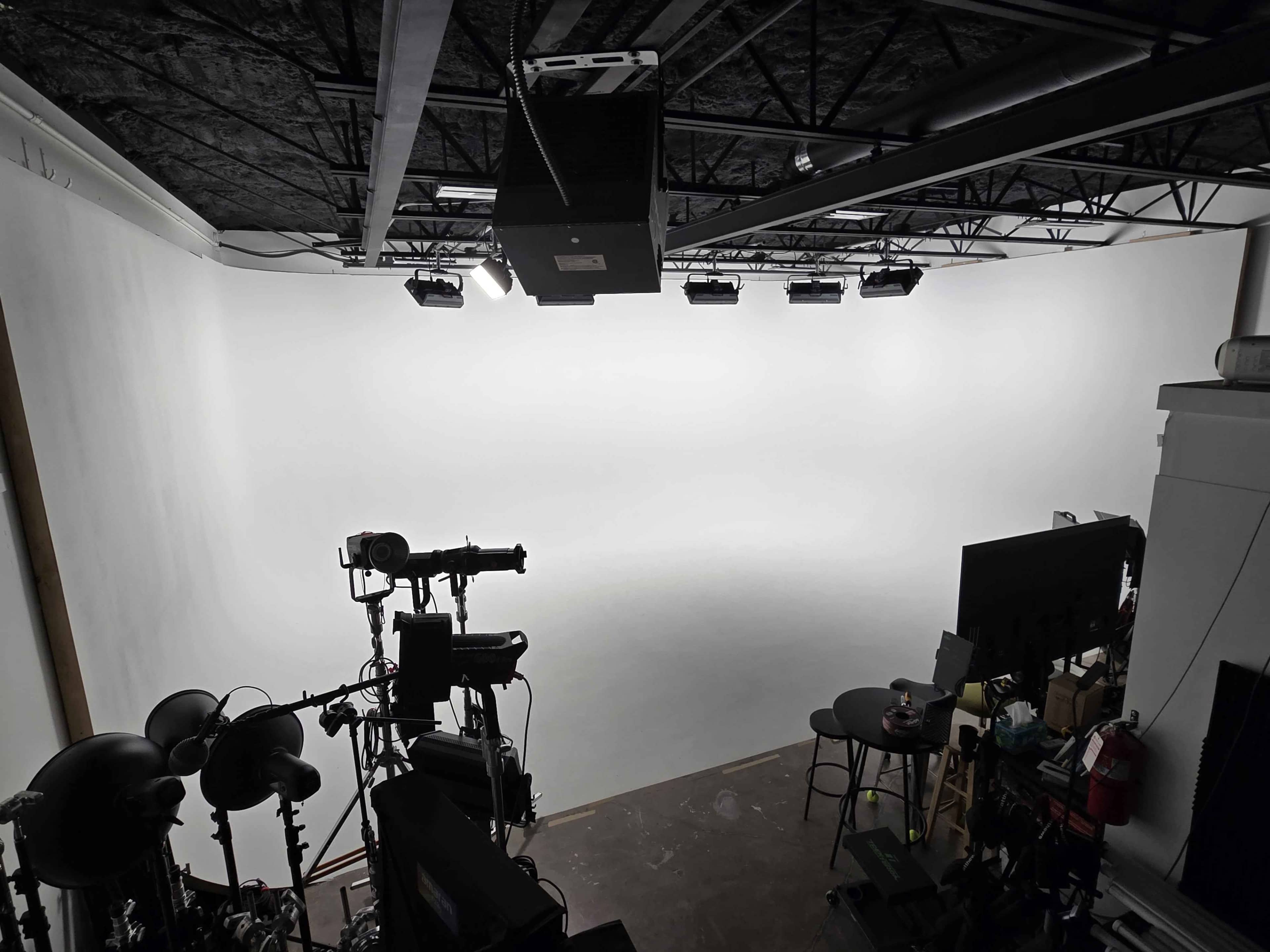 A photography studio features a spacious white backdrop, camera equipment, and simple furniture set against an industrial ceiling.