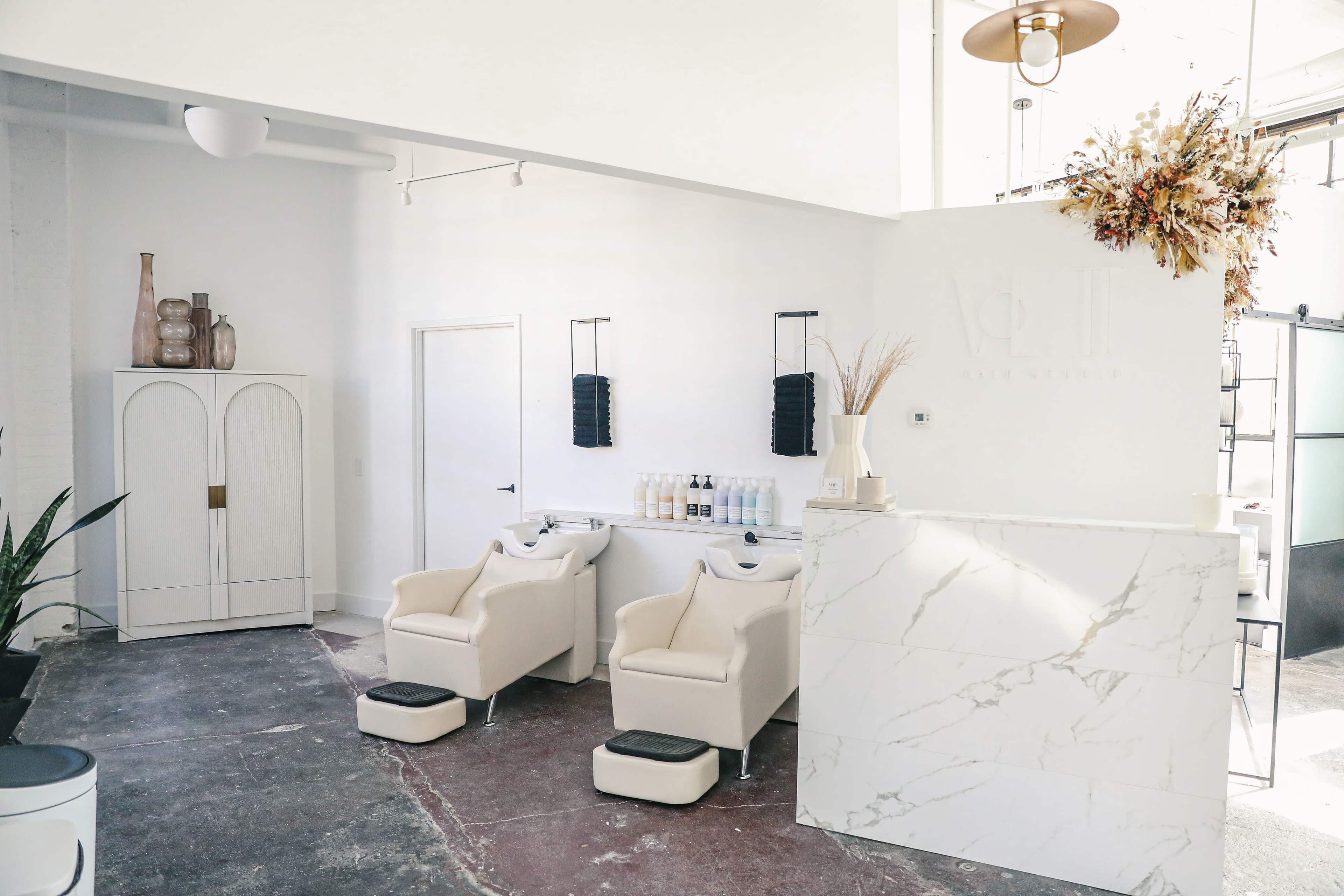 The interior of a modern hair salon features two styling chairs, a reception desk made of white marble, and minimalist decor with a large white cabinet and decorative vases.