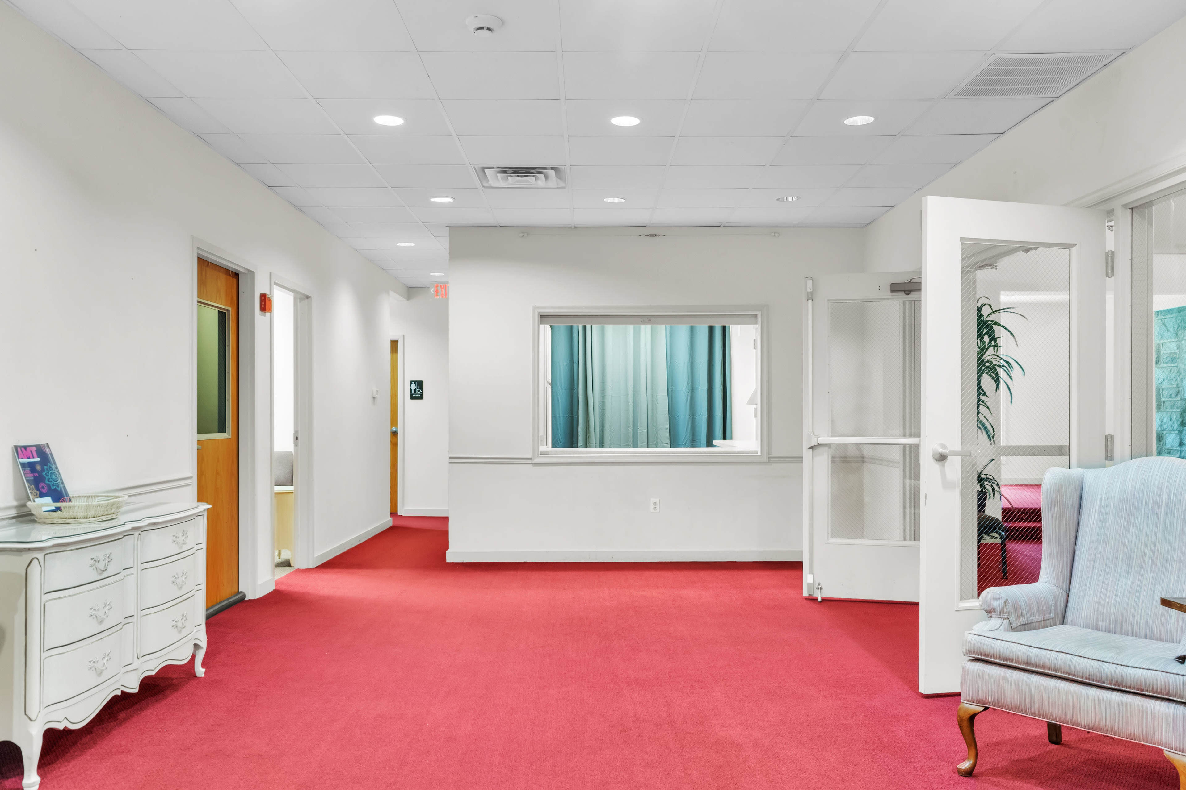 A corridor with red carpeting features several doors, a window partition, and a decorative white console table.