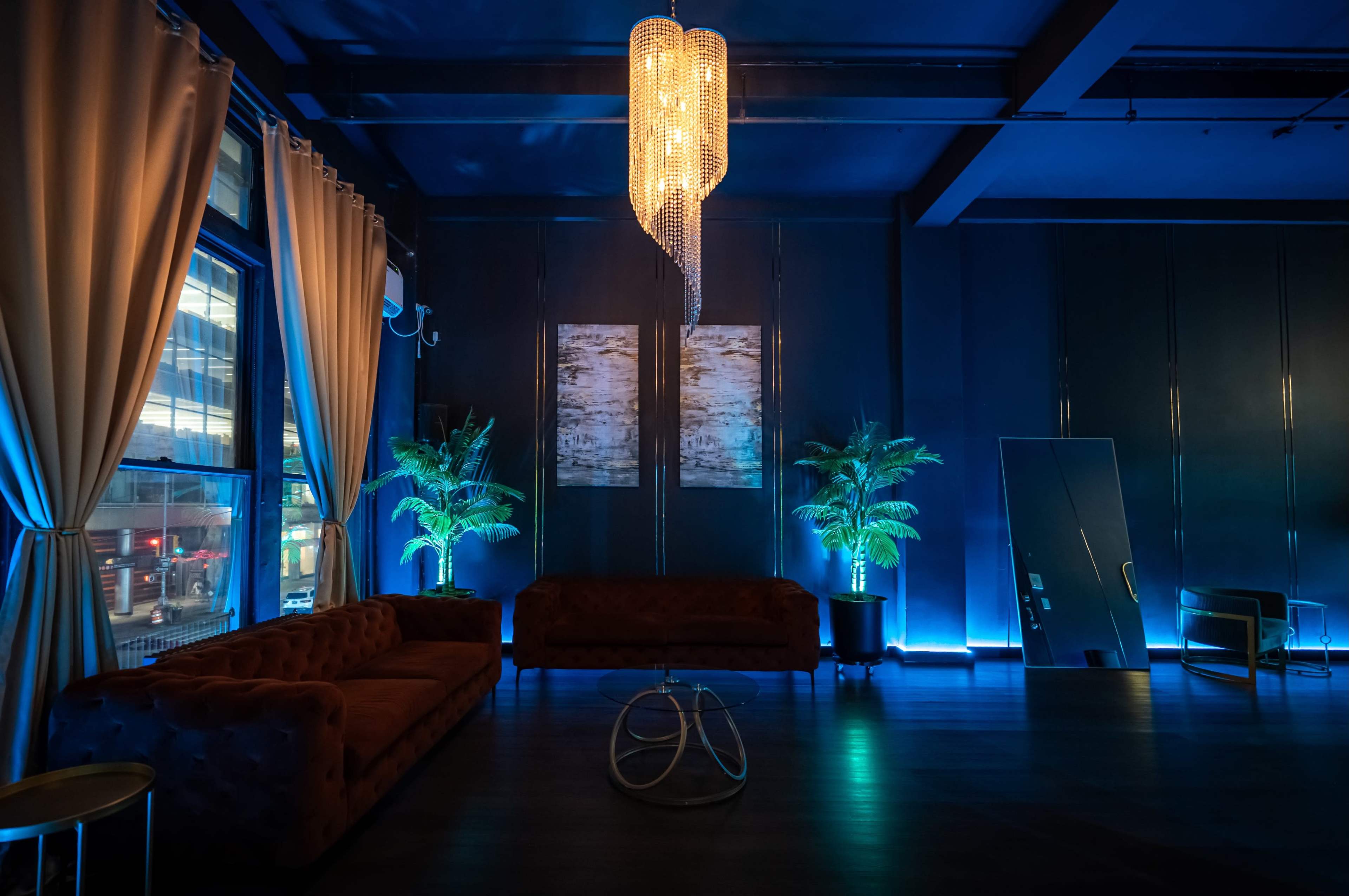 The image shows a dimly lit lounge area featuring two brown sofas, a tall chandelier, decorative plants, and a large mirror against a blue-lit wall.