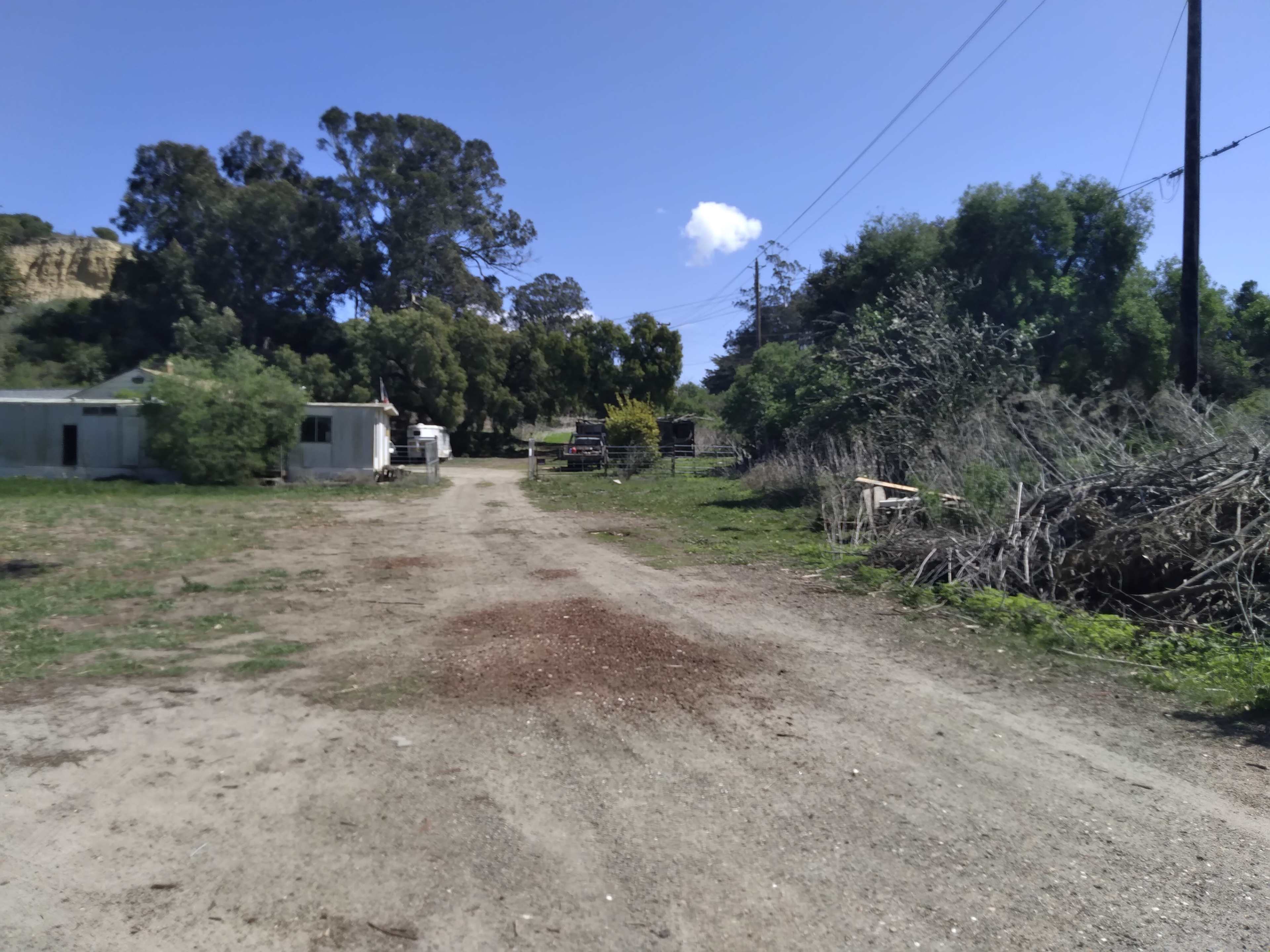 A dirt road leads through a rural area with sparse buildings on one side and greenery on the other.