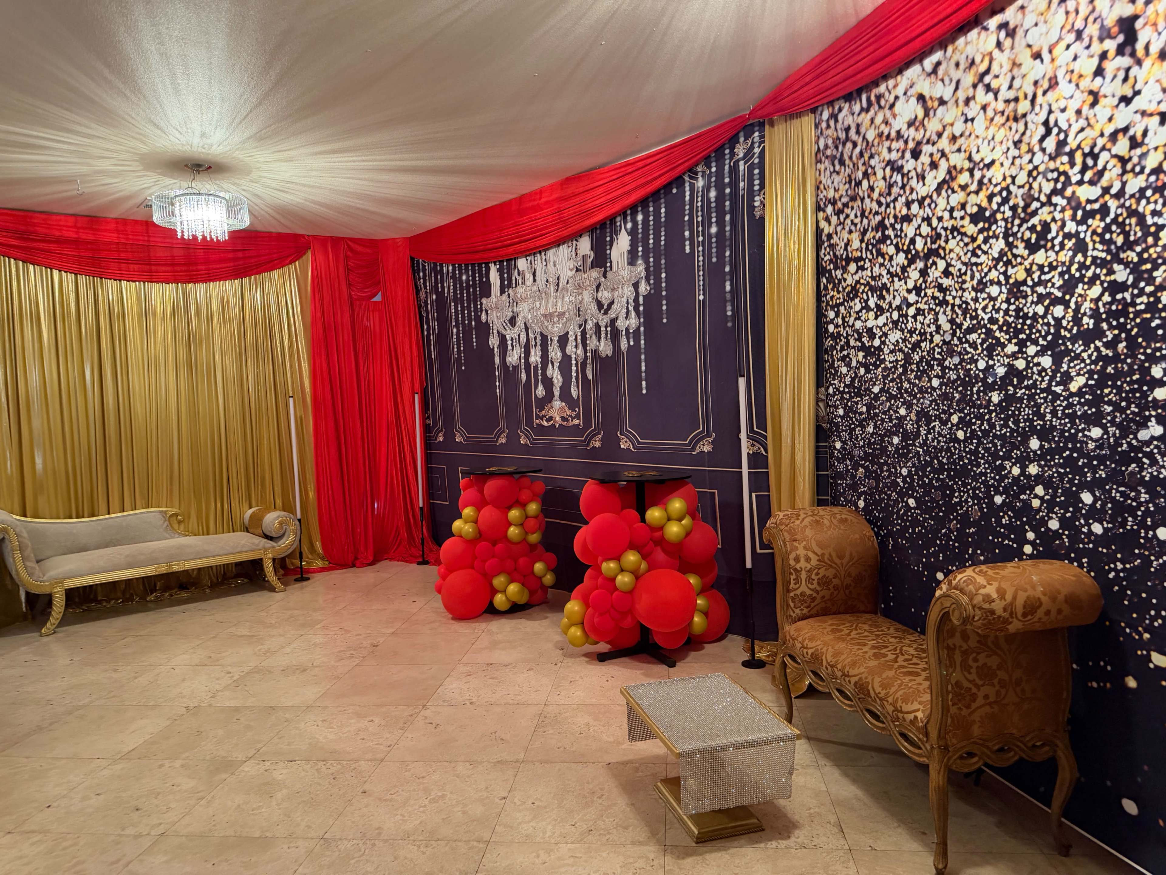 The image features a decorated event space with gold and red drapes, a chandelier, and two ornate chairs flanking a display of red and yellow balloons.