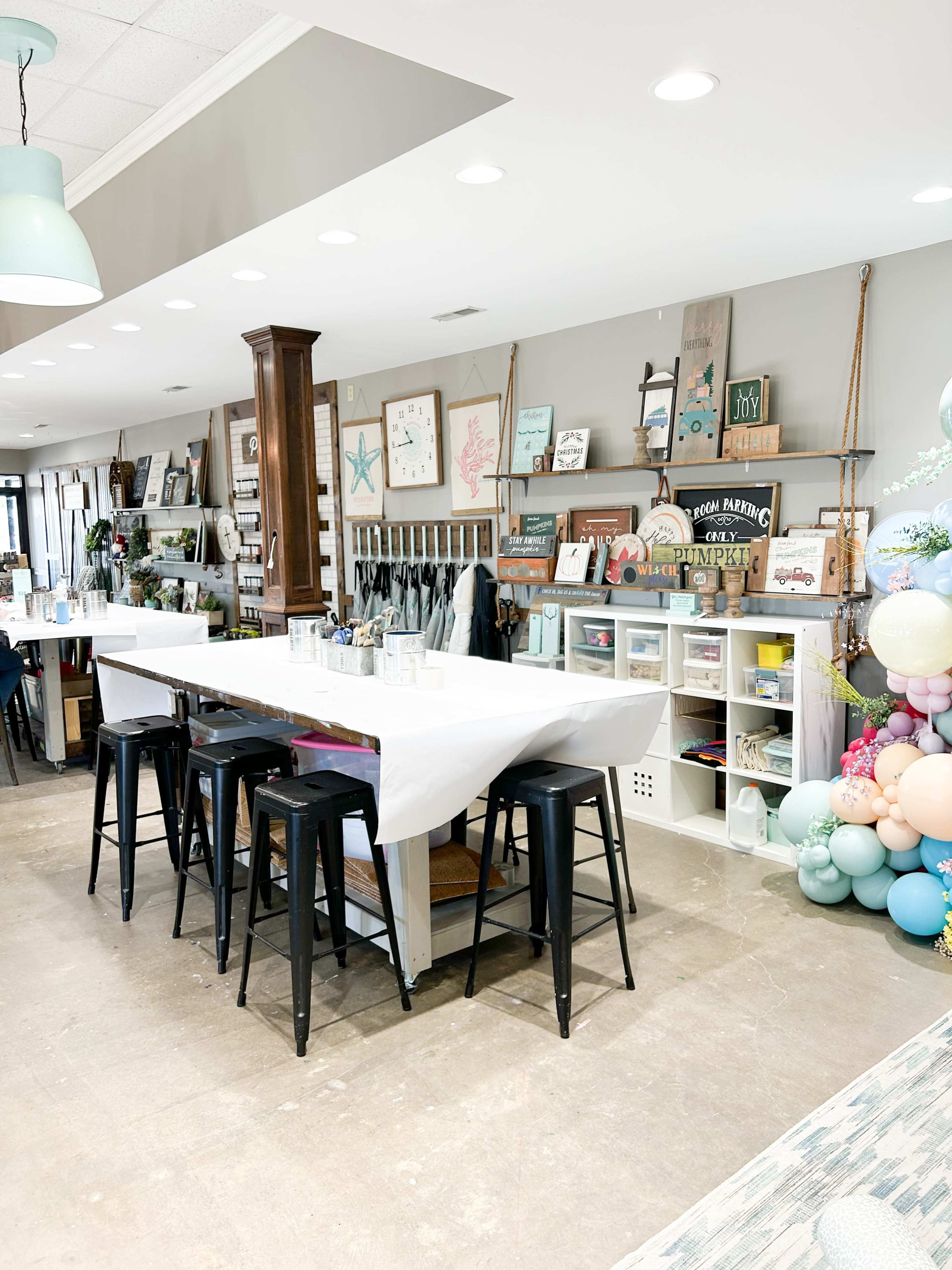 A spacious room features a long table with stools, surrounded by shelves displaying various decorative items and artwork.