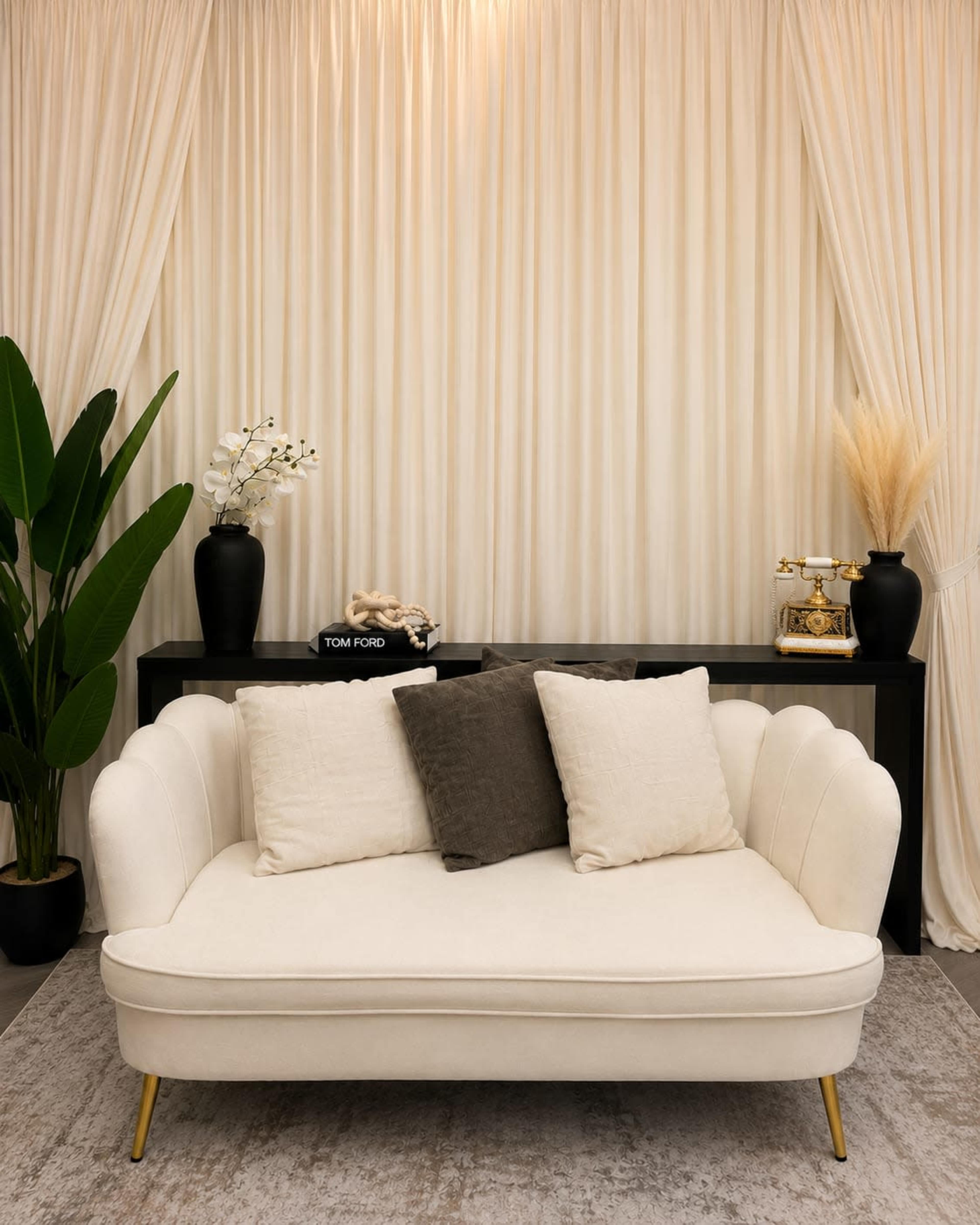 A white two-seater couch with three cushions is positioned in front of a black console table, adorned with decorative plants and vases, against a backdrop of sheer curtains.