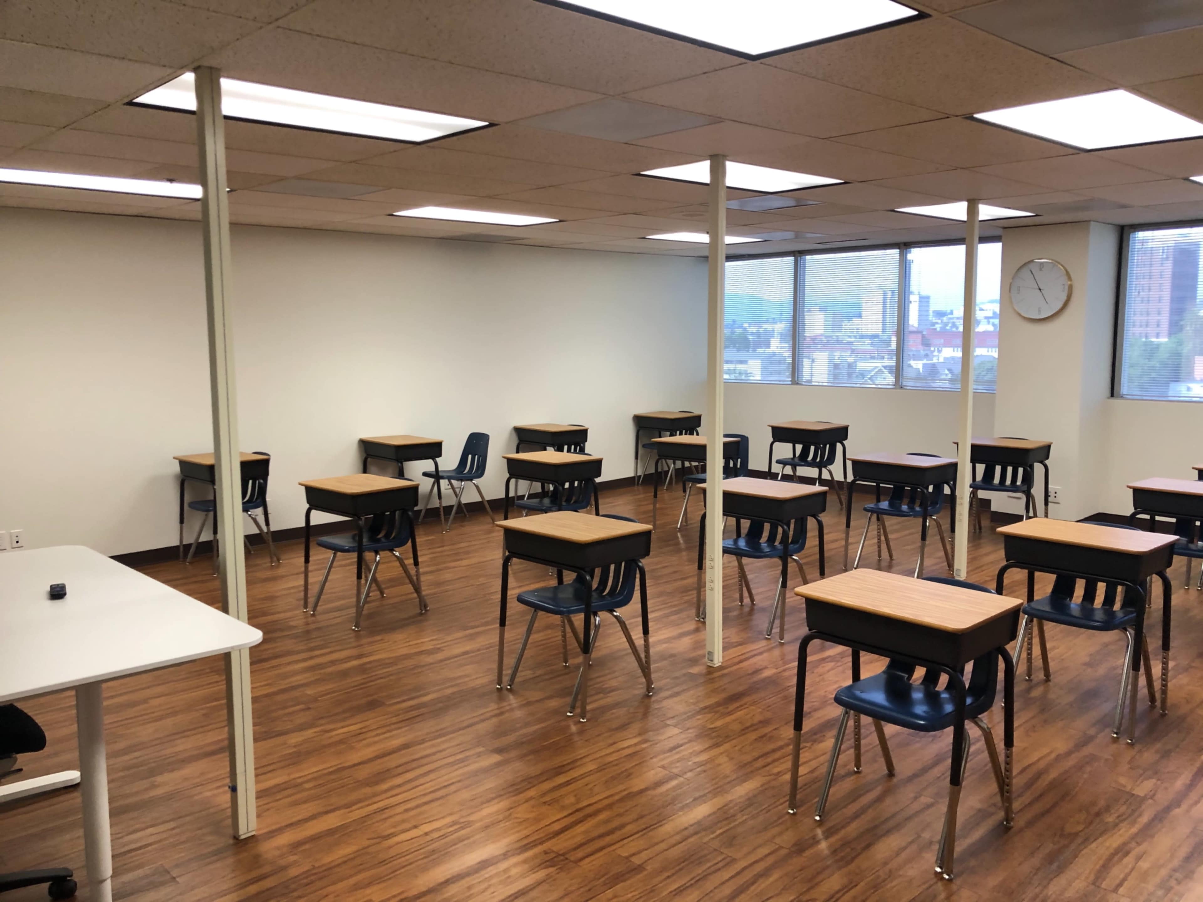 The image shows a classroom with several wooden desks and metal chairs arranged in a spacious room with large windows and a clock on the wall.