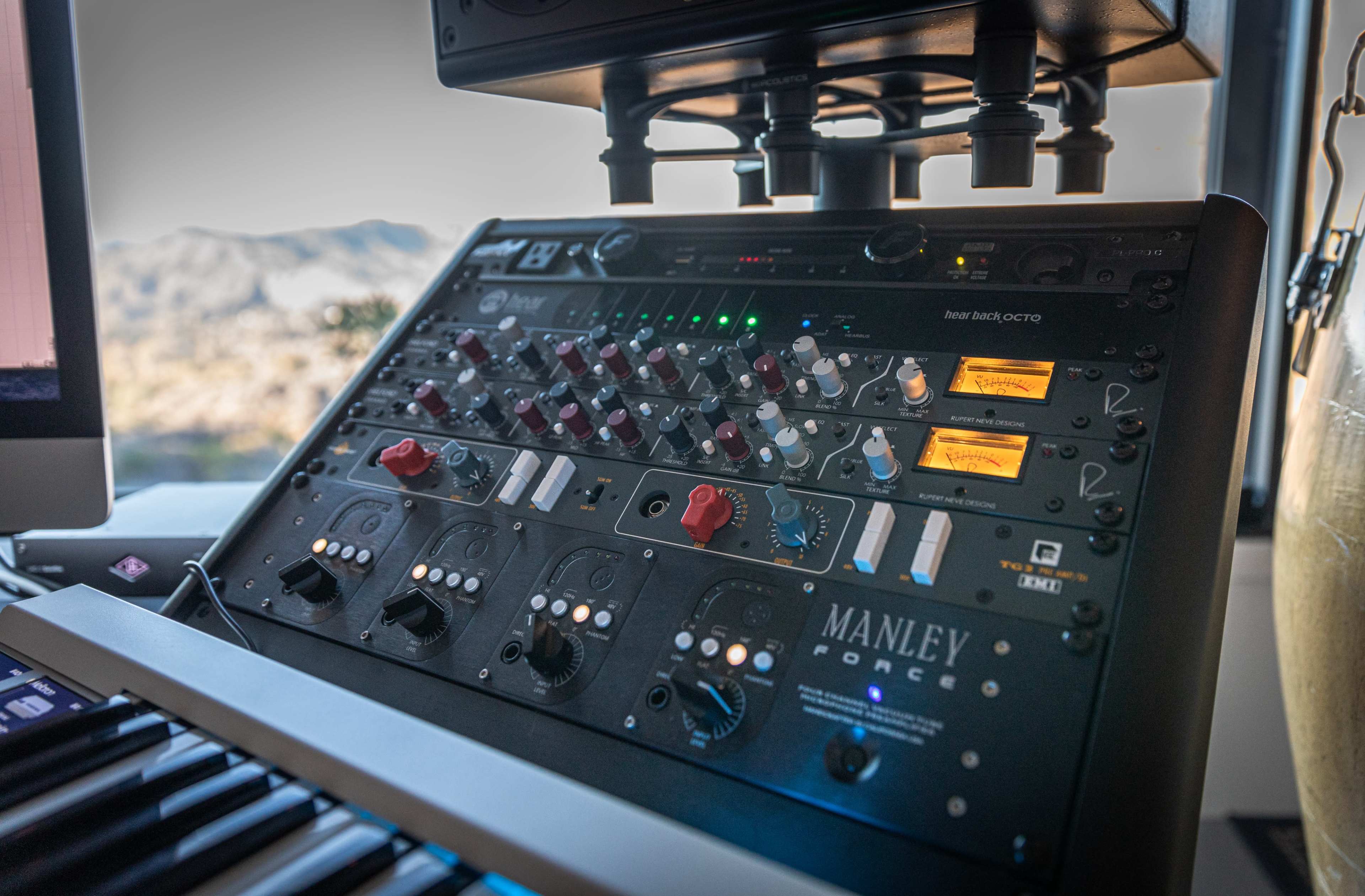 A close-up of a Manley audio mixing console with various knobs, sliders, and illuminated indicators, positioned on a desk next to a computer and keyboard.