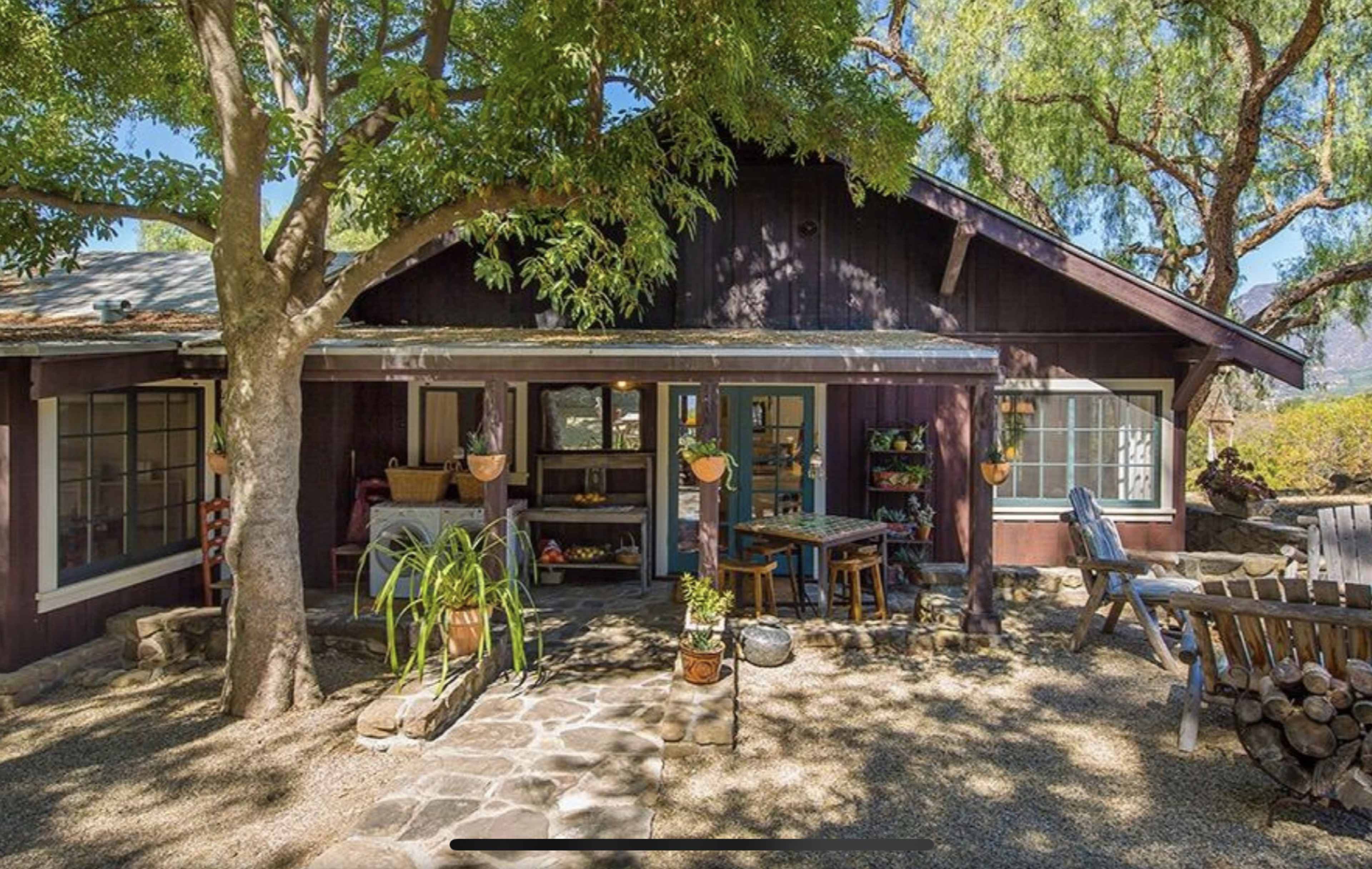 A rustic, wooden house surrounded by trees and a stone pathway features an outdoor sitting area with garden decorations and a table.