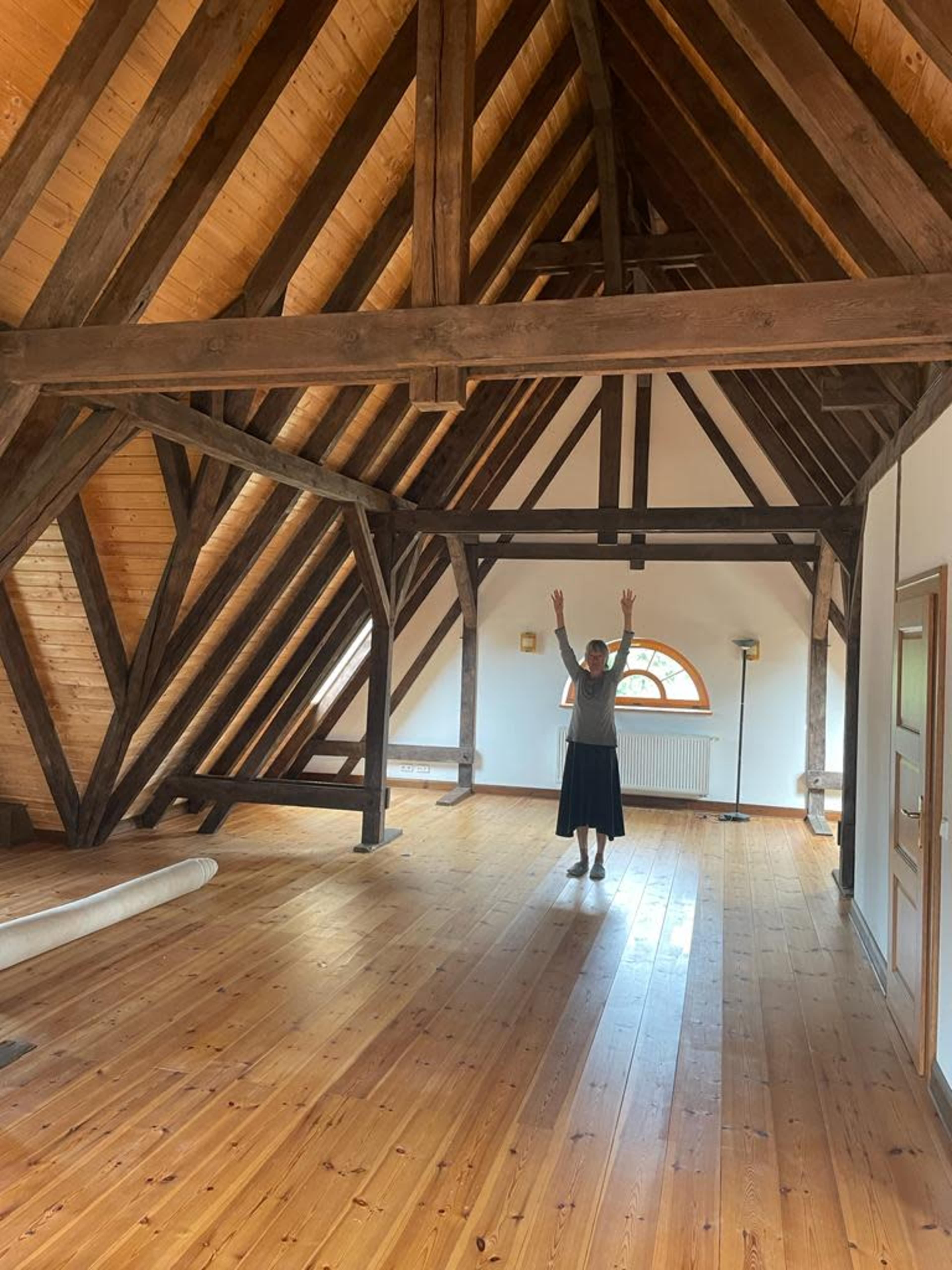 A person stands with arms raised in the center of a spacious room with wooden beams and a hardwood floor.