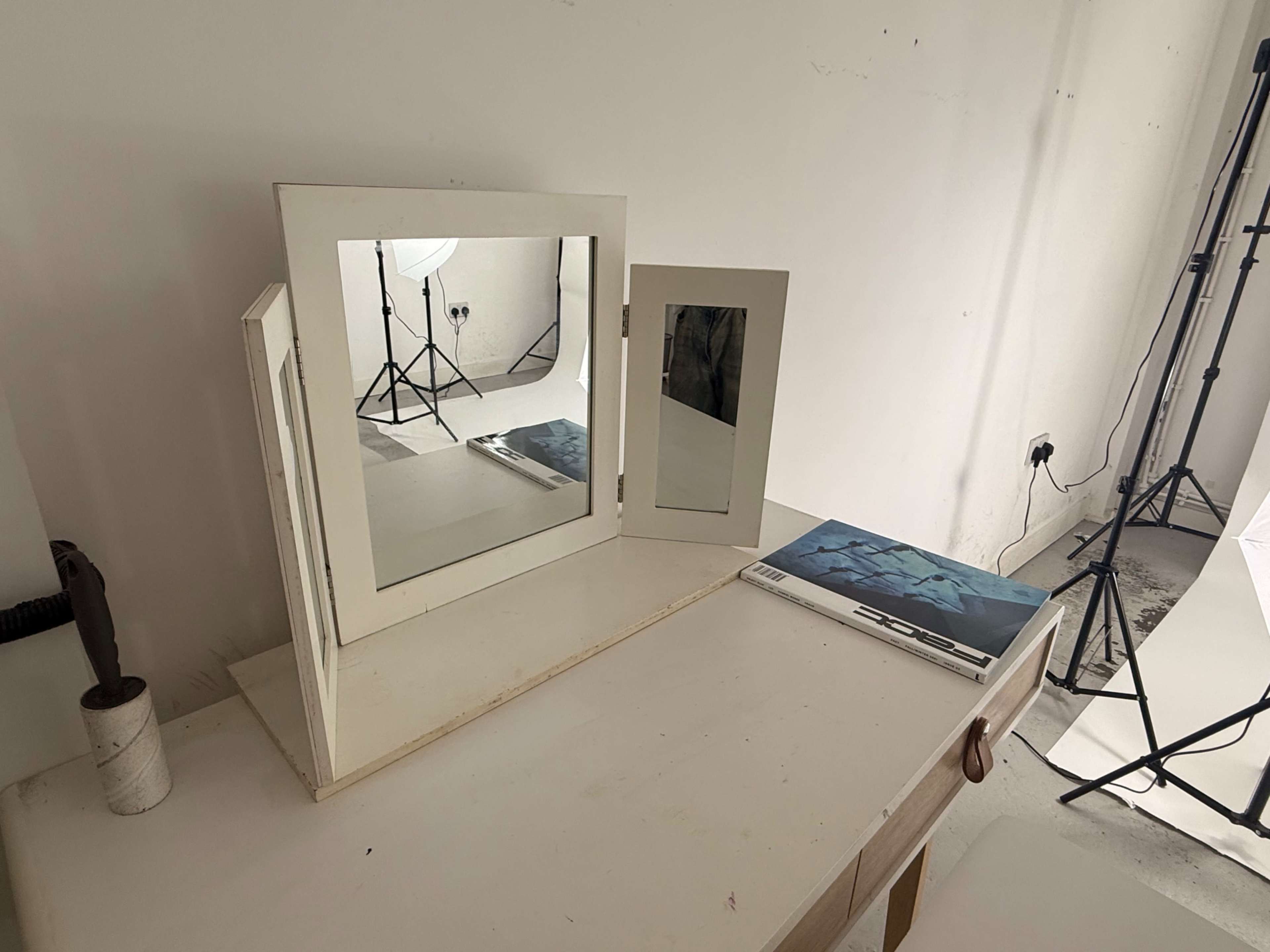 A three-panel mirror is placed on a table next to a photography backdrop, with a book lying open on the surface.