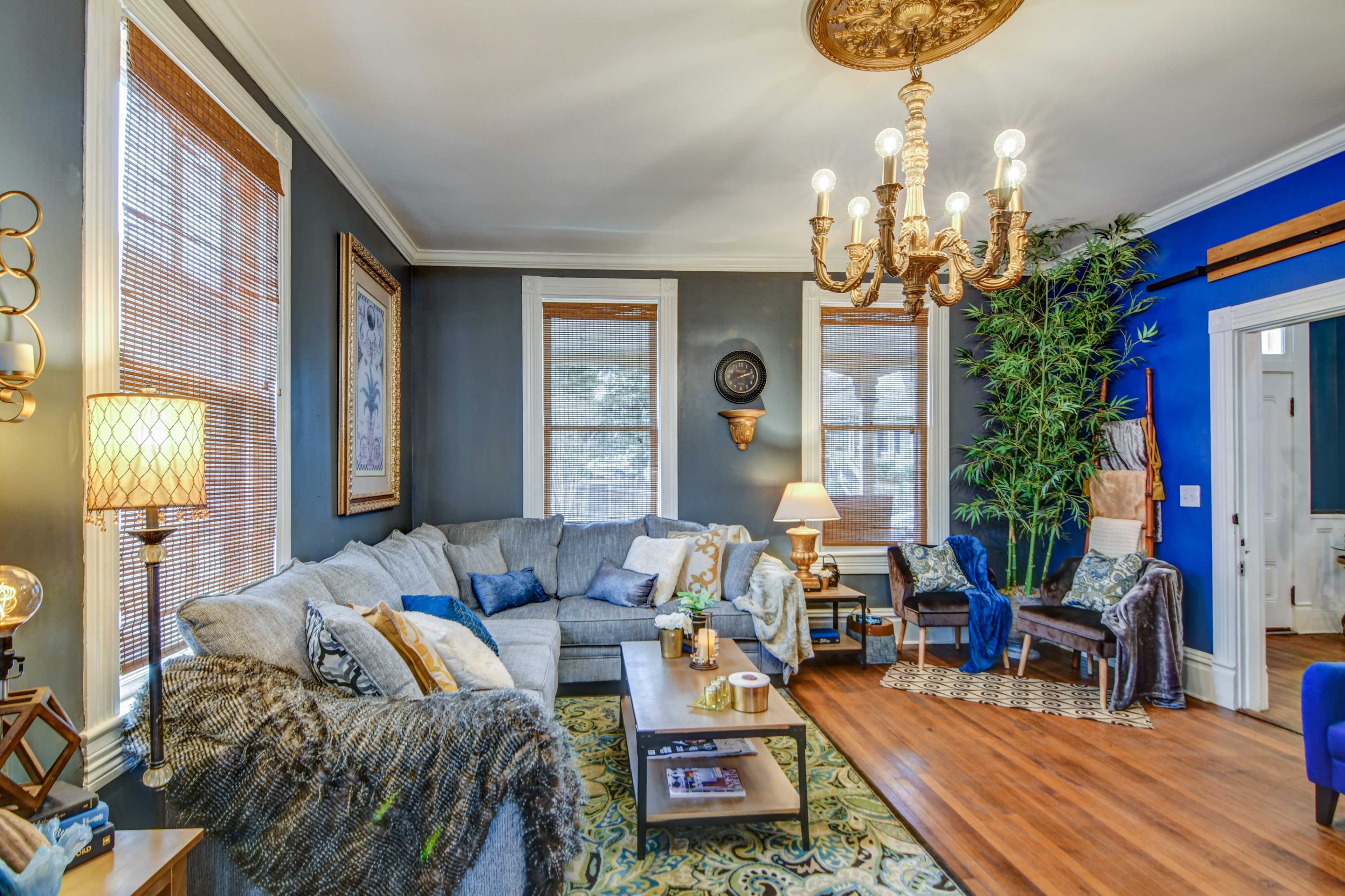A spacious living room features a large gray sofa, wooden coffee table, and a chandelier, with blue and neutral walls and natural light from the windows.