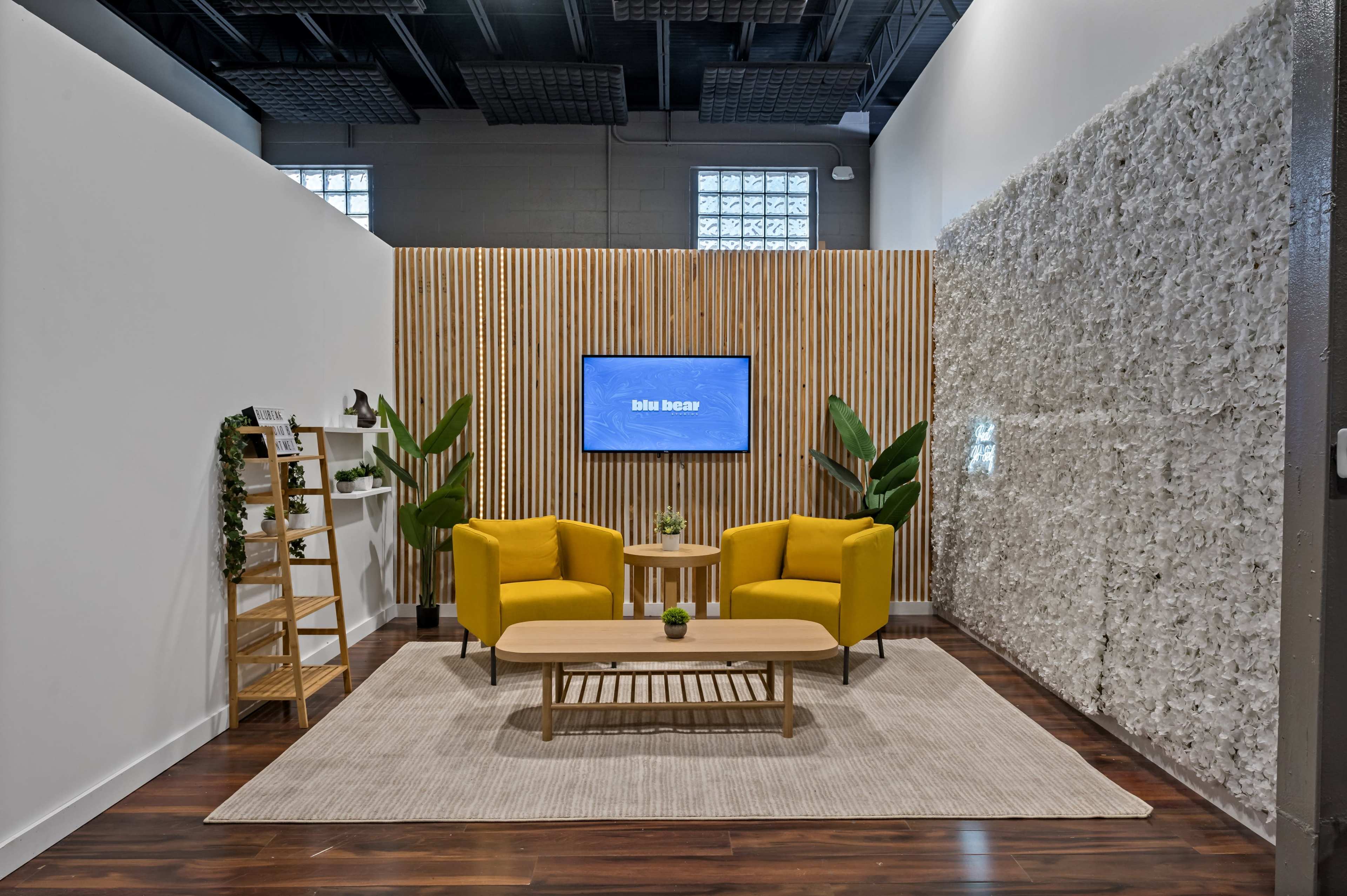 A modern seating area features two yellow chairs and a wooden table on a rug, flanked by a decorative wall and indoor plants, with a television mounted on a wooden backdrop.