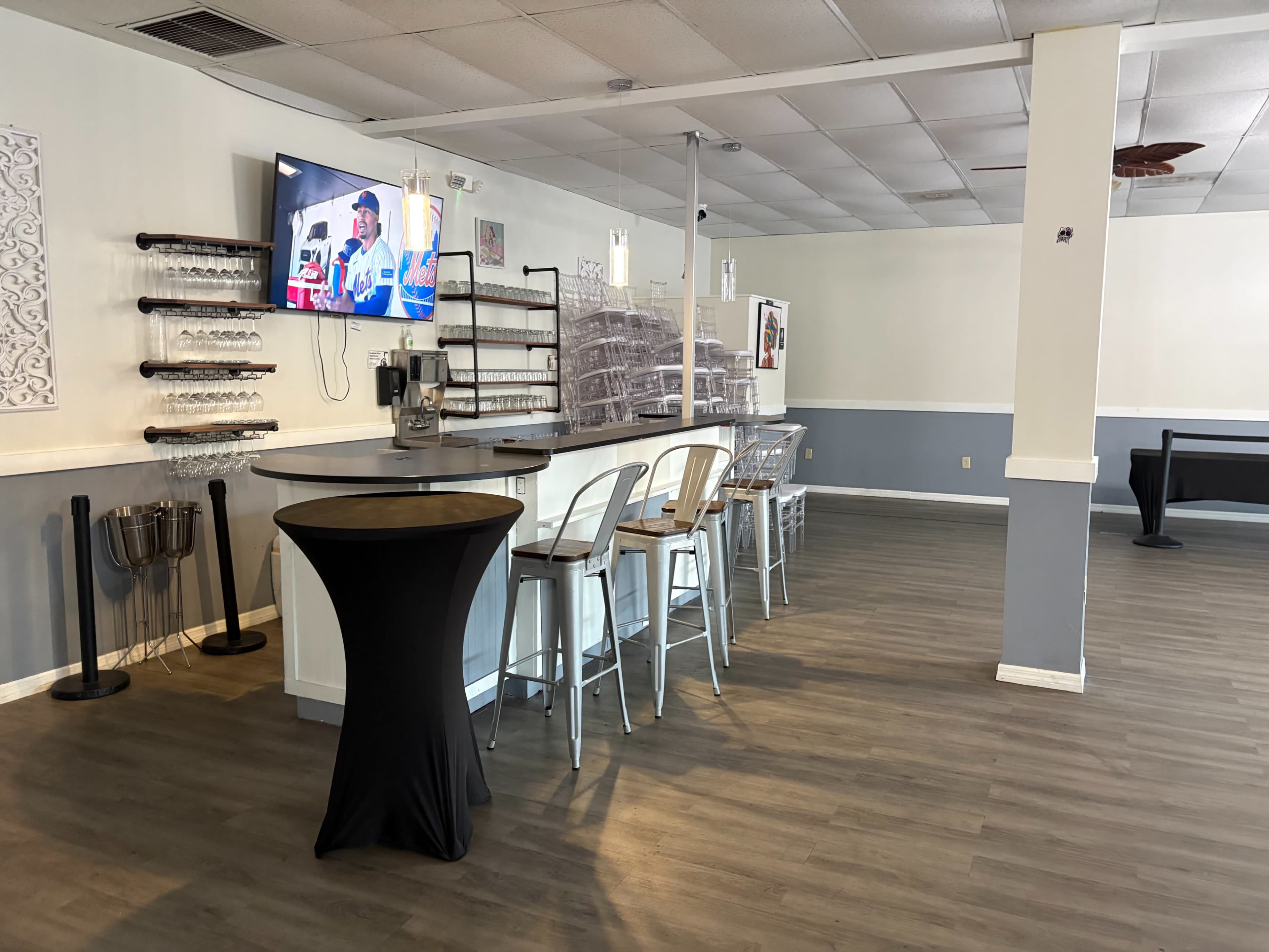 The image shows a spacious interior of a bar or restaurant, featuring a counter with stools, a television mounted on the wall, and a mix of seating arrangements including a tall table with chairs.