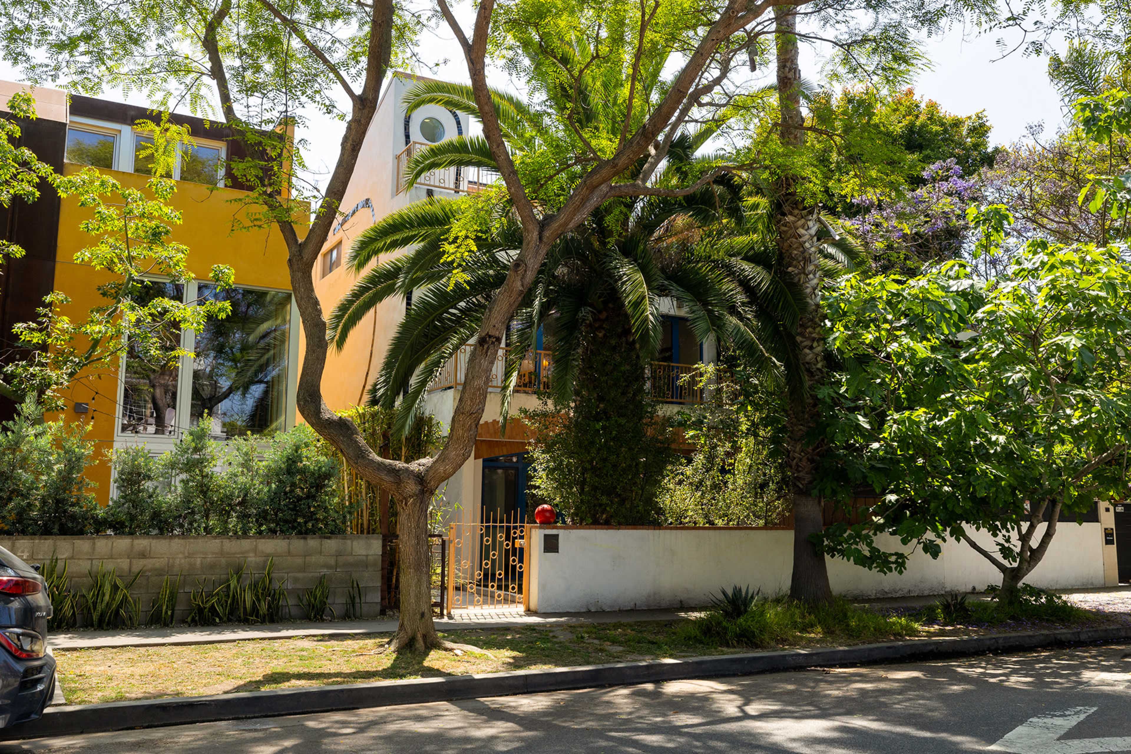 The image shows a colorful two-story house surrounded by lush trees and shrubs on a sunny street.