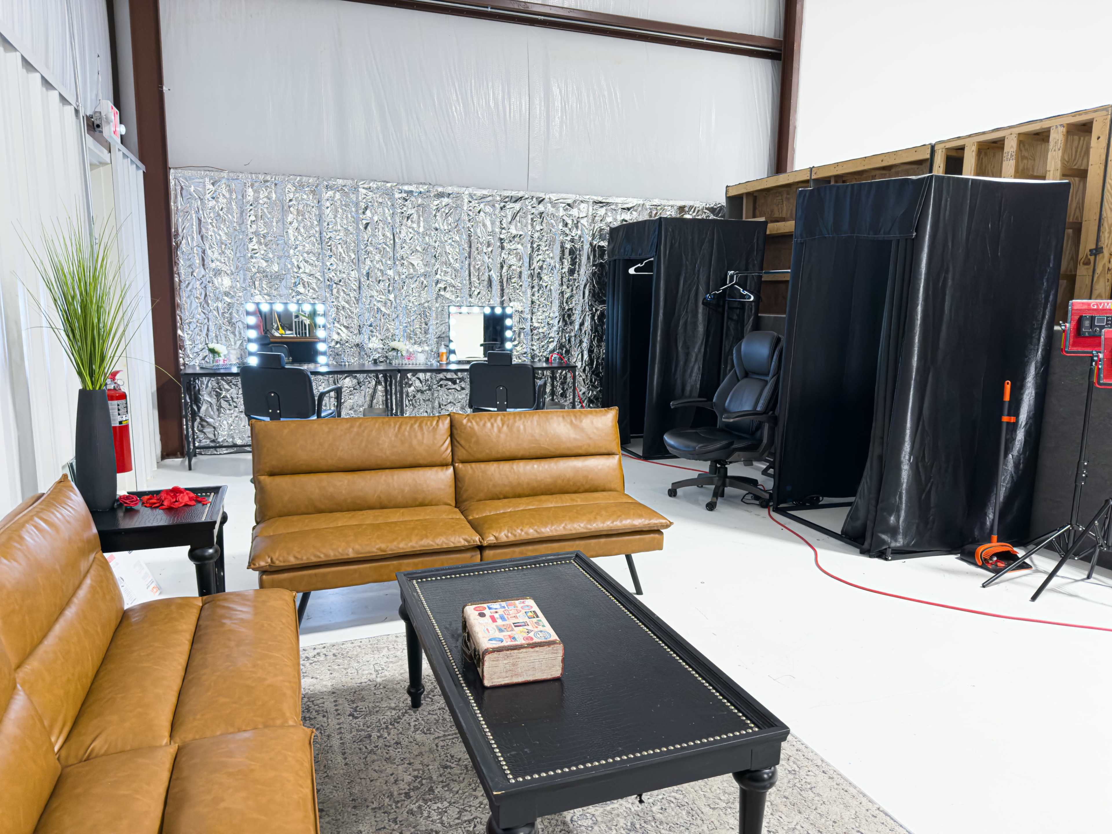 The image shows a spacious room with brown leather sofas, a black chair, a table in the background with vanity mirrors, and a sectioned-off area with black curtains.