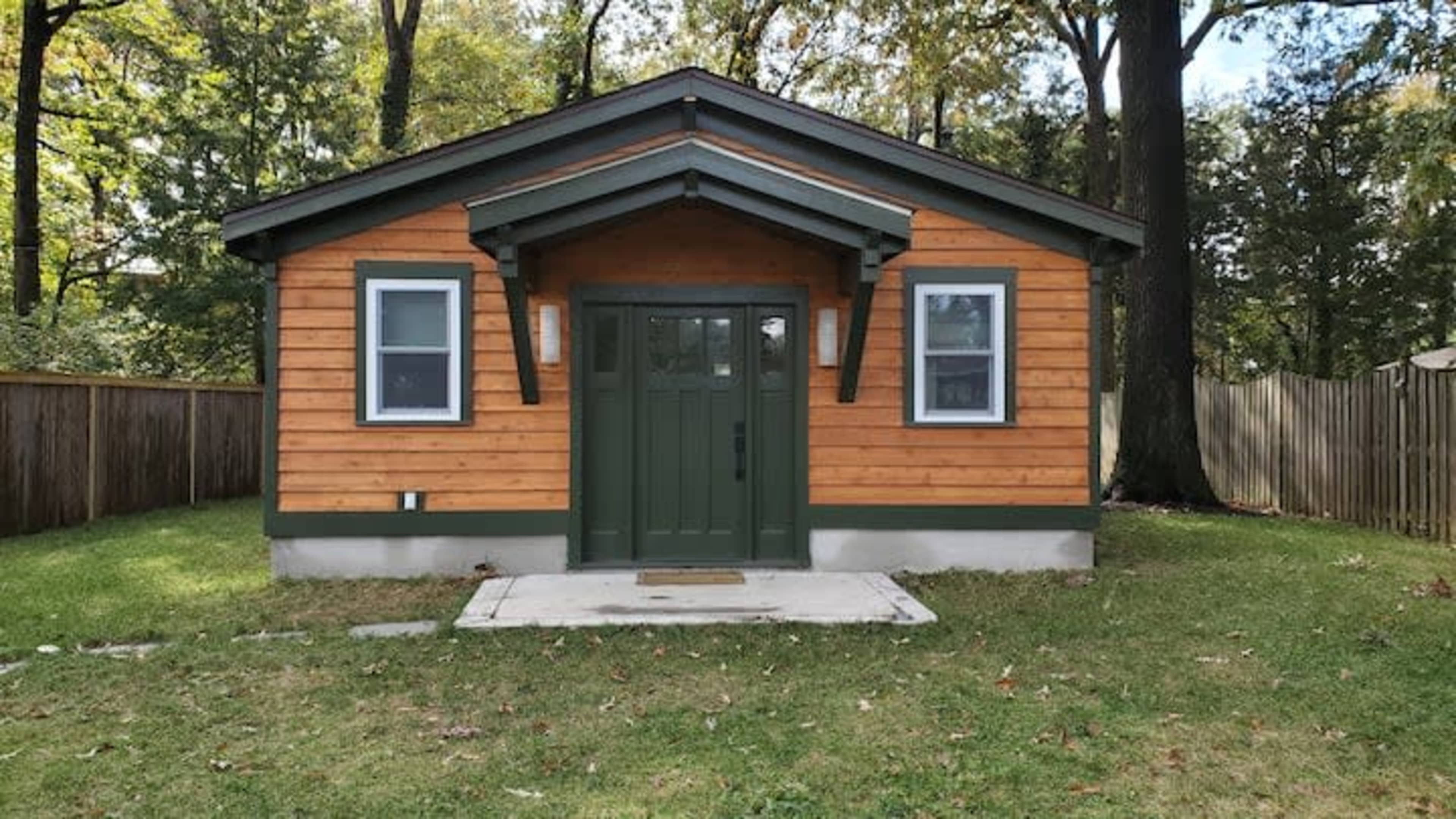 The image shows a small, single-story house with a wooden exterior and a dark green front door surrounded by well-maintained landscaping.