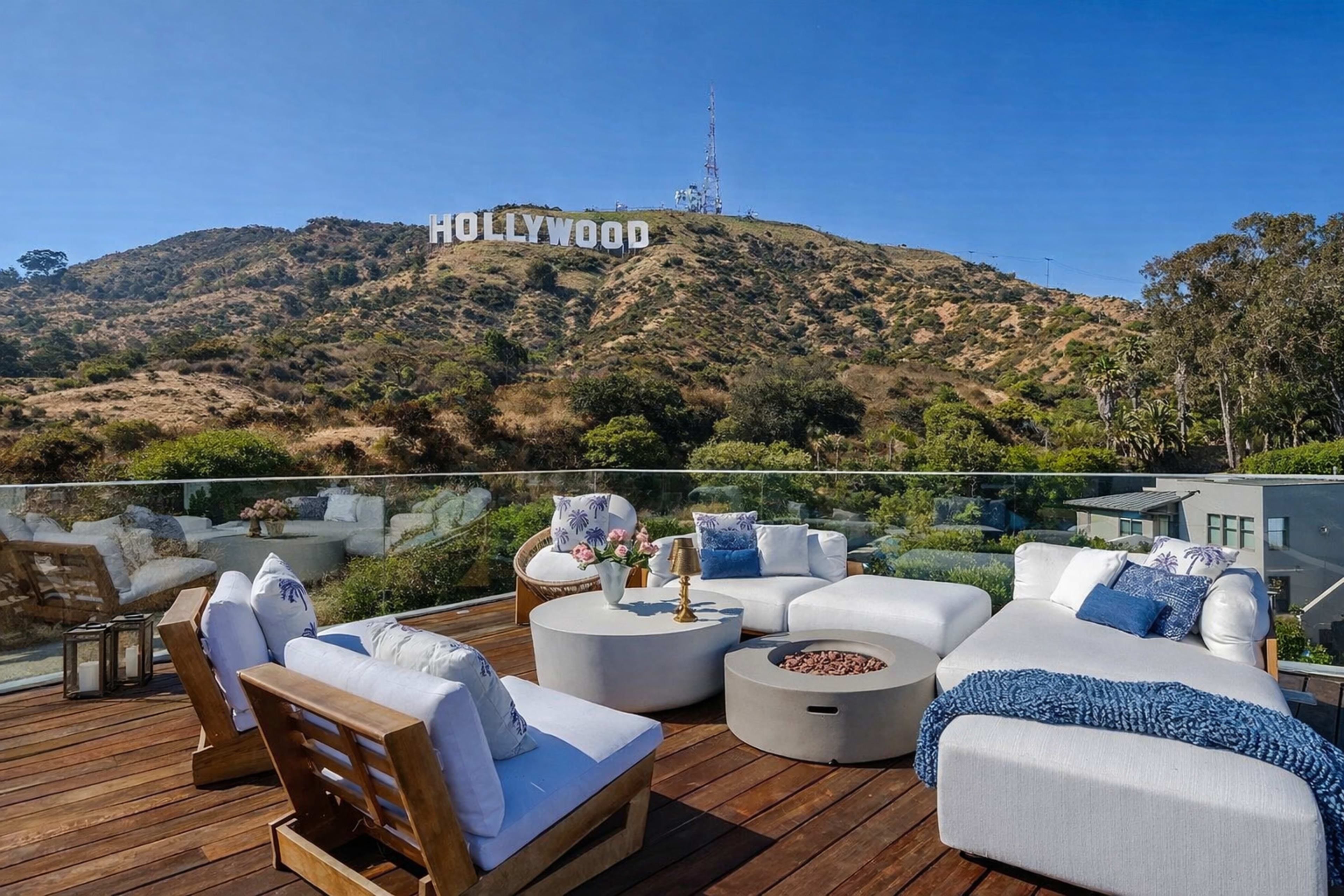 A terrace with modern furniture overlooks the Hollywood sign on a hillside in bright sunshine.