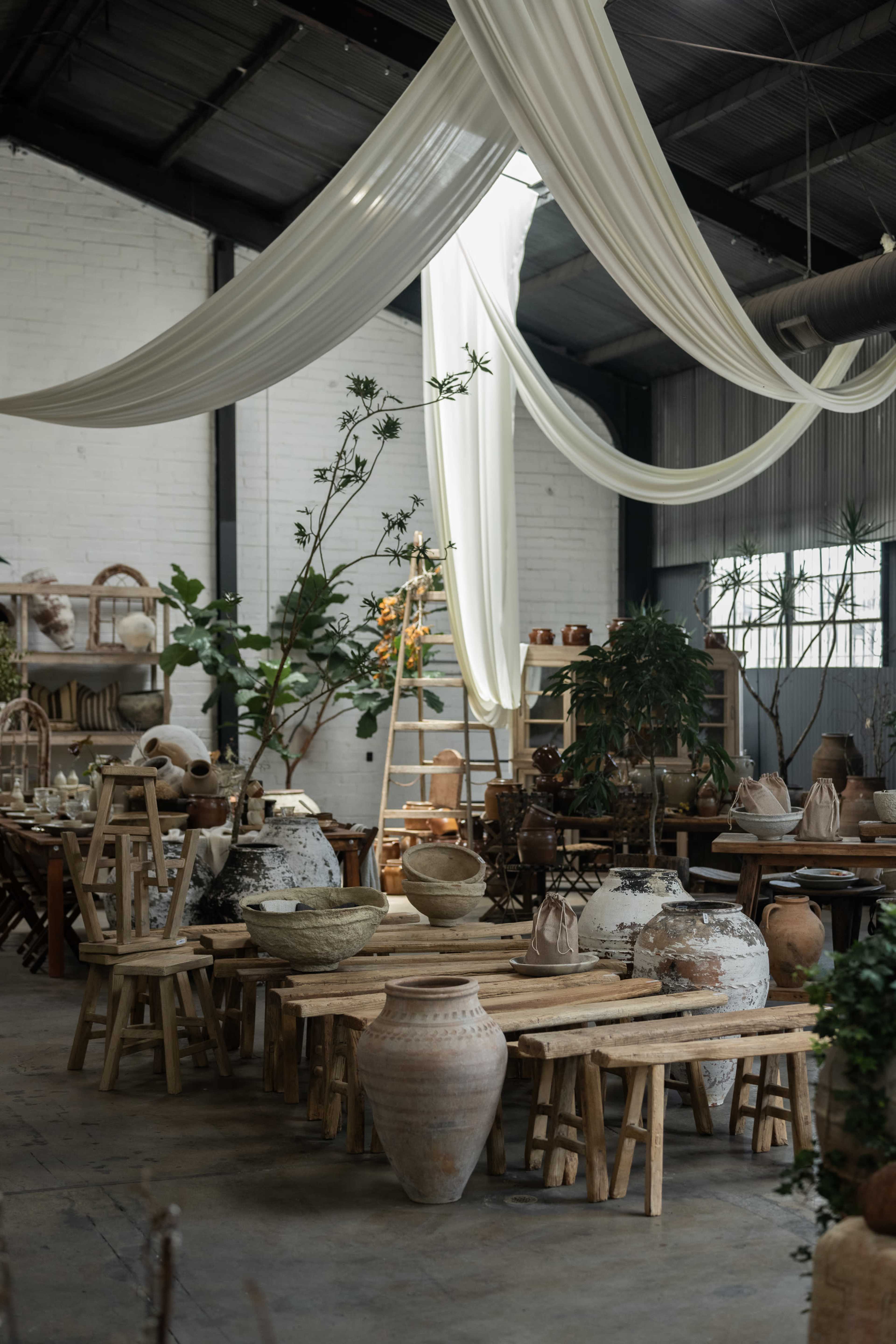 A spacious interior features wooden tables adorned with various pottery pieces, with greenery and draped fabric hanging from the ceiling.