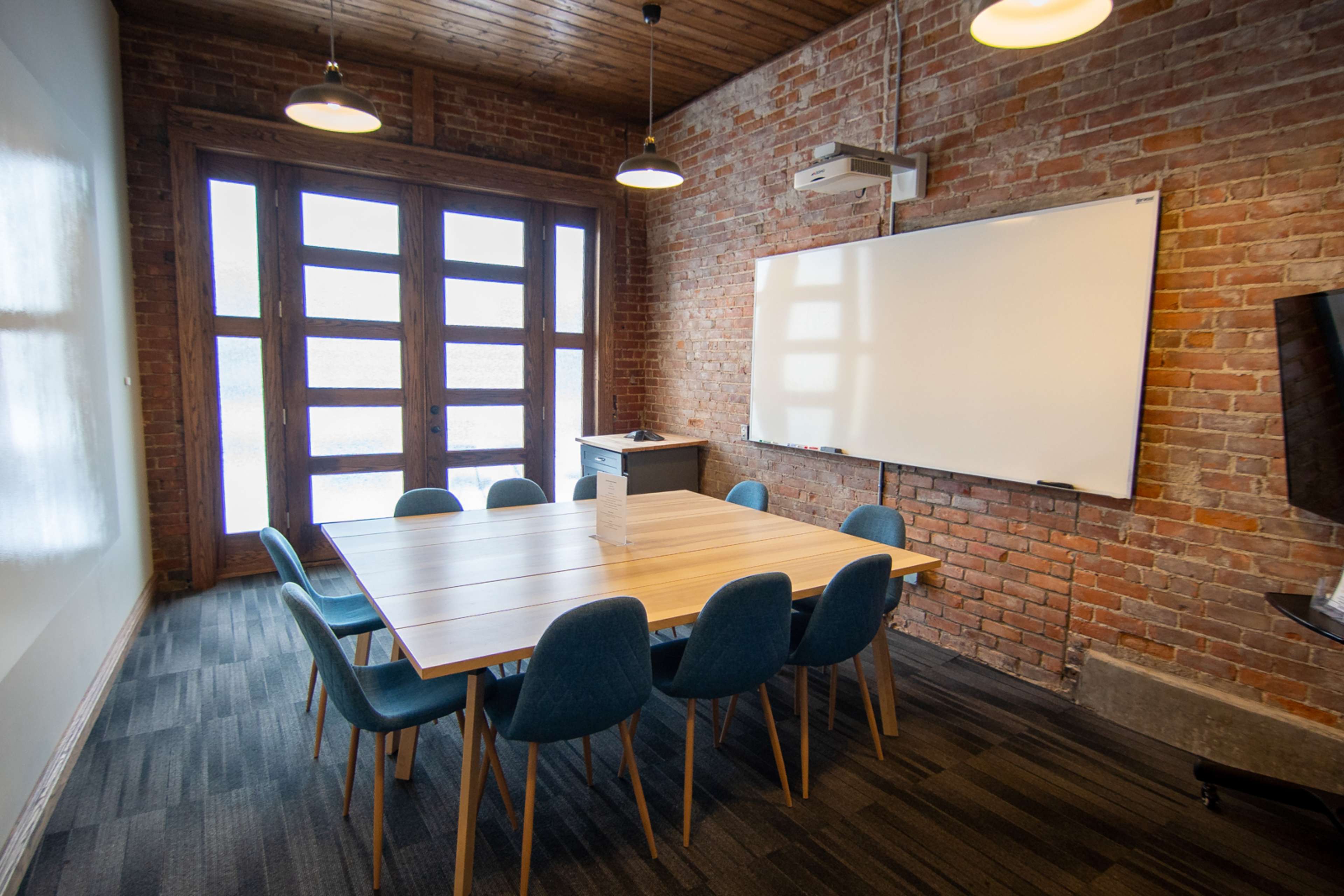 A conference room with a large wooden table surrounded by blue chairs, exposed brick walls, and a whiteboard on one side.