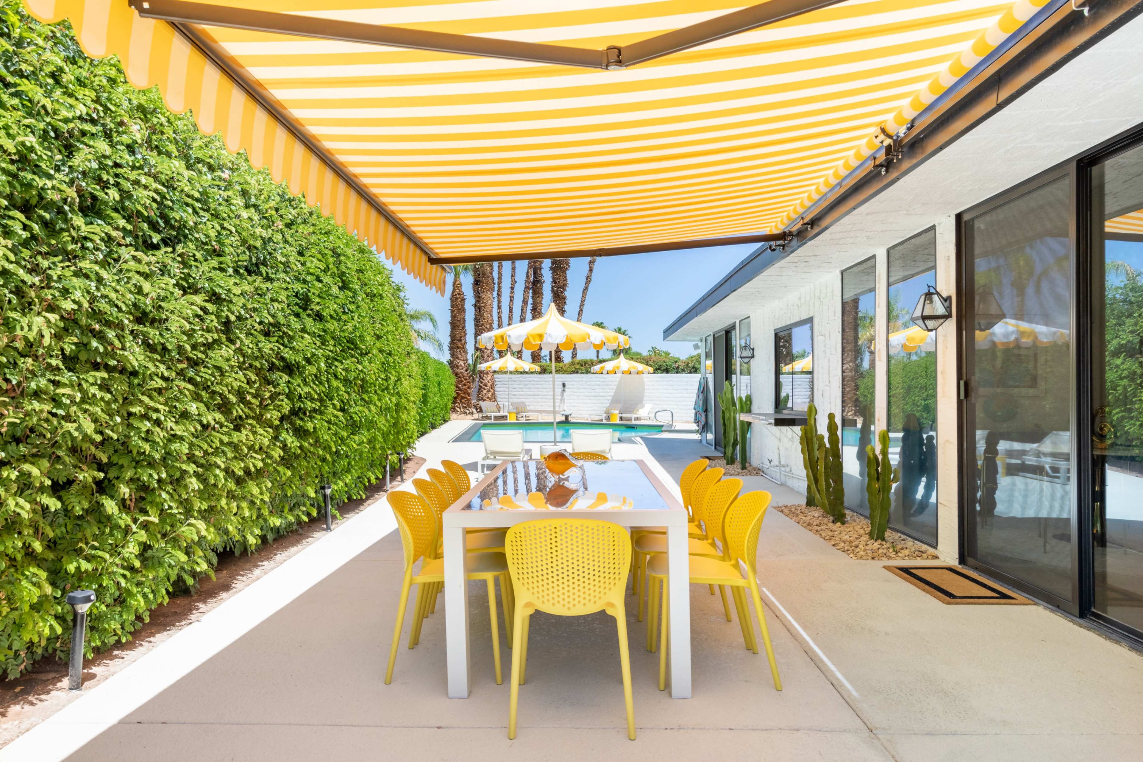 A patio area features a large dining table surrounded by yellow chairs, shaded by a striped yellow awning, with a pool and landscaped greenery in the background.