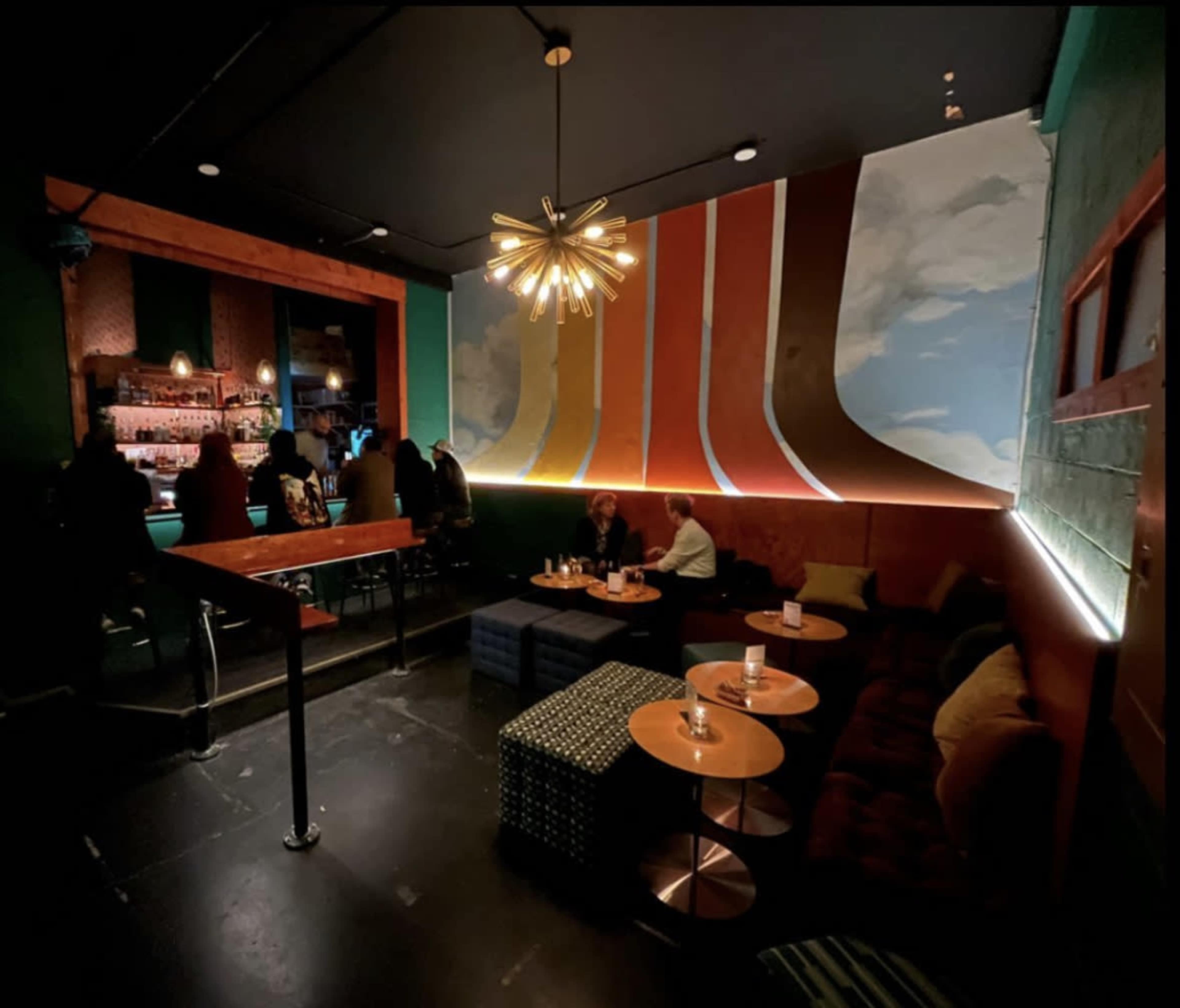 A dimly lit bar features colorful wall art, cozy seating, and patrons gathered around small tables.