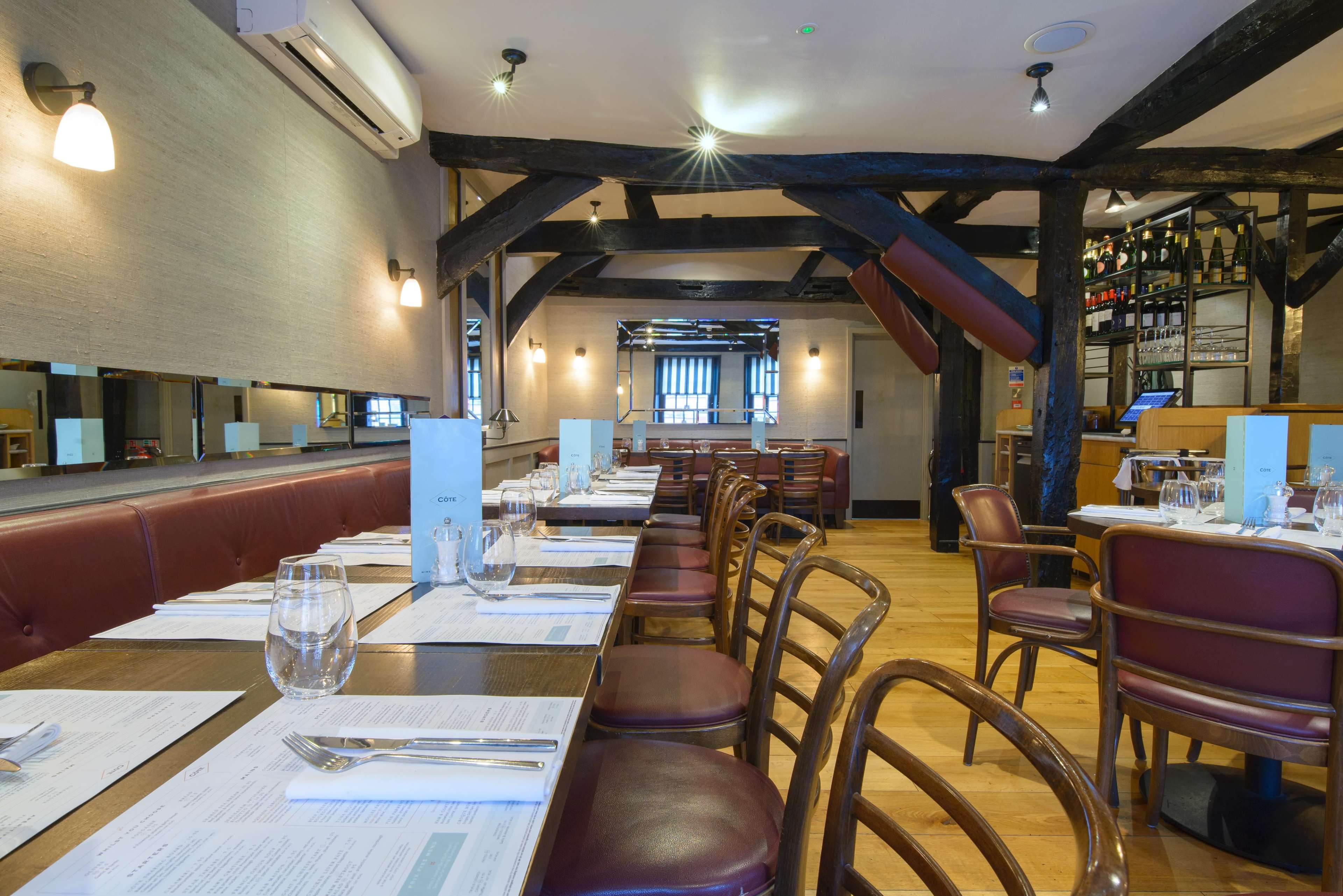 The image shows an interior view of a restaurant featuring wooden beams, red upholstered seating, and neatly arranged tables with glassware and menus.