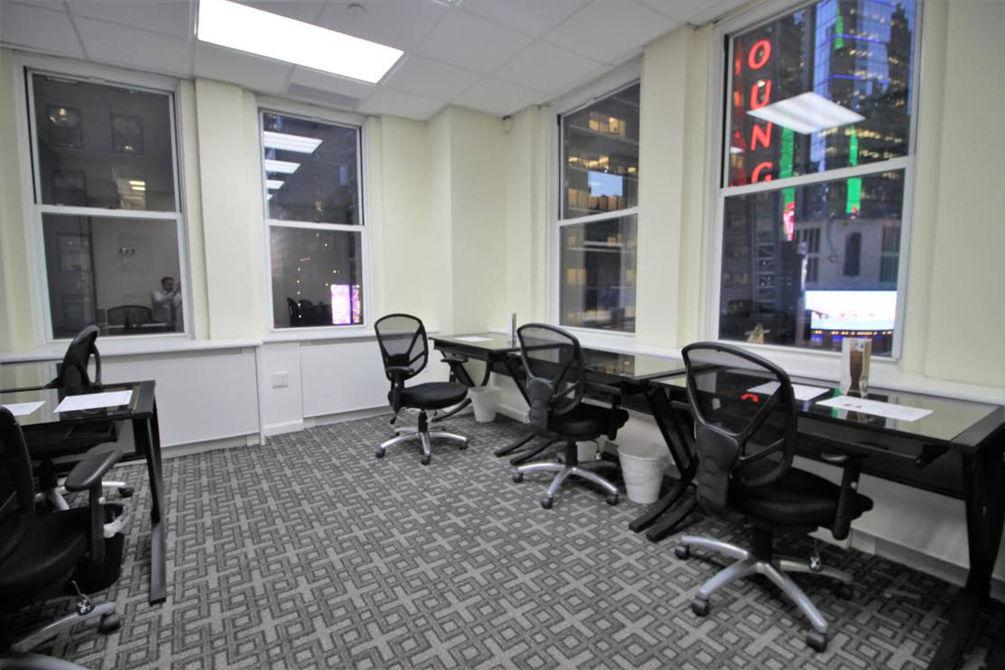 The image shows an office space with several empty desks and chairs, illuminated by natural light from large windows.