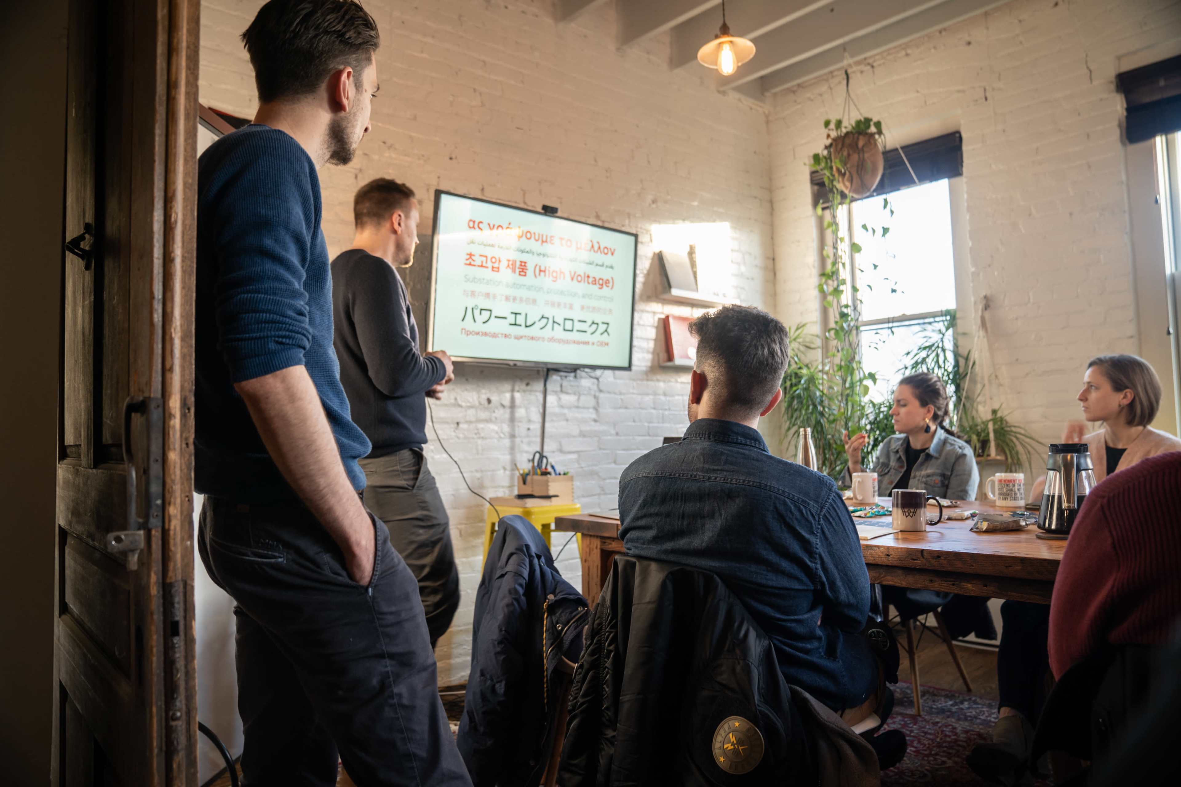 A group of five people is seated around a wooden table in a bright room, while one person stands and points at a presentation displayed on a wall-mounted screen.