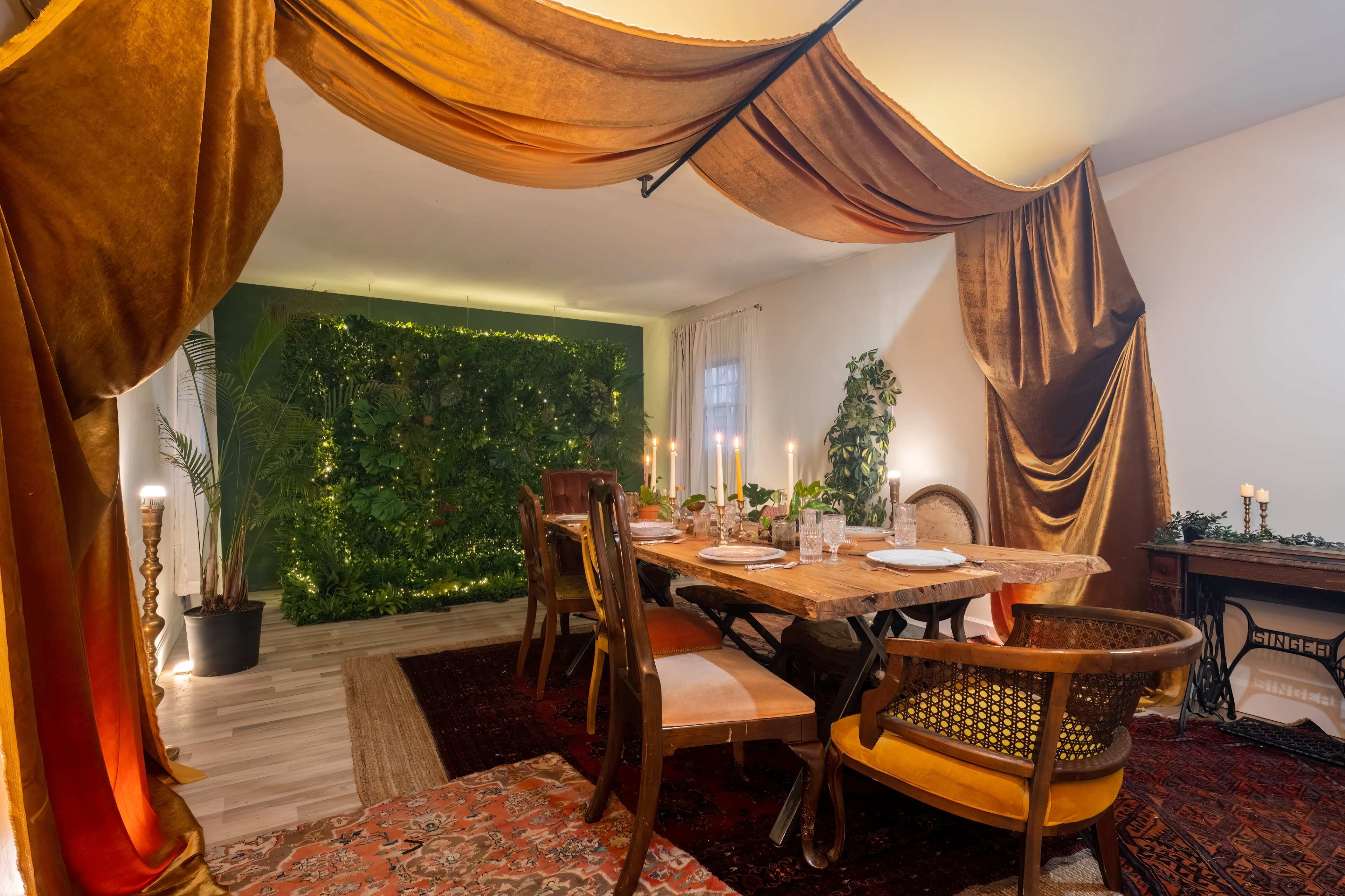 A dining area features a wooden table set with dishes, surrounded by chairs and draped fabric, with a backdrop of greenery and ambient lighting.