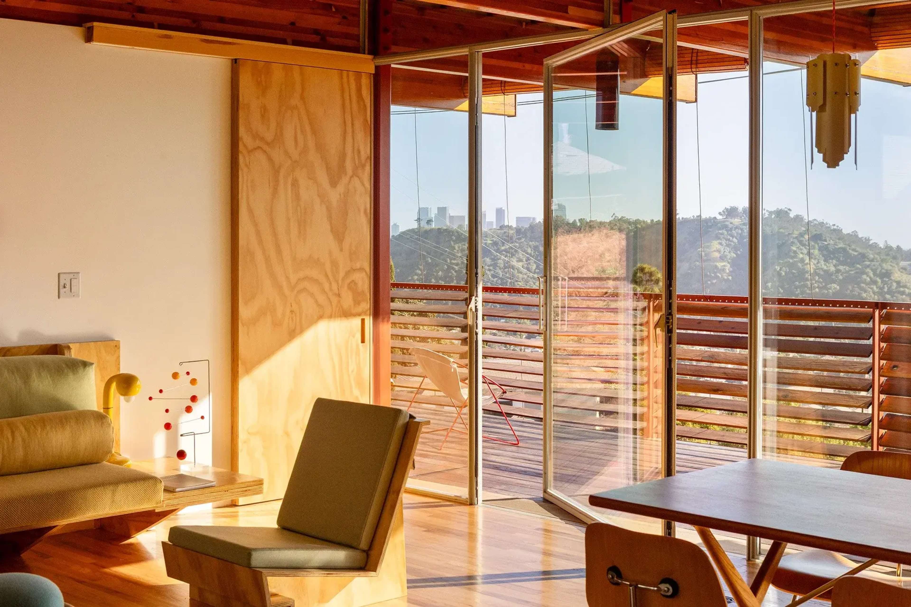 The image shows a modern interior space featuring wooden elements, large glass doors, and a view of a mountainous landscape with city buildings in the distance.