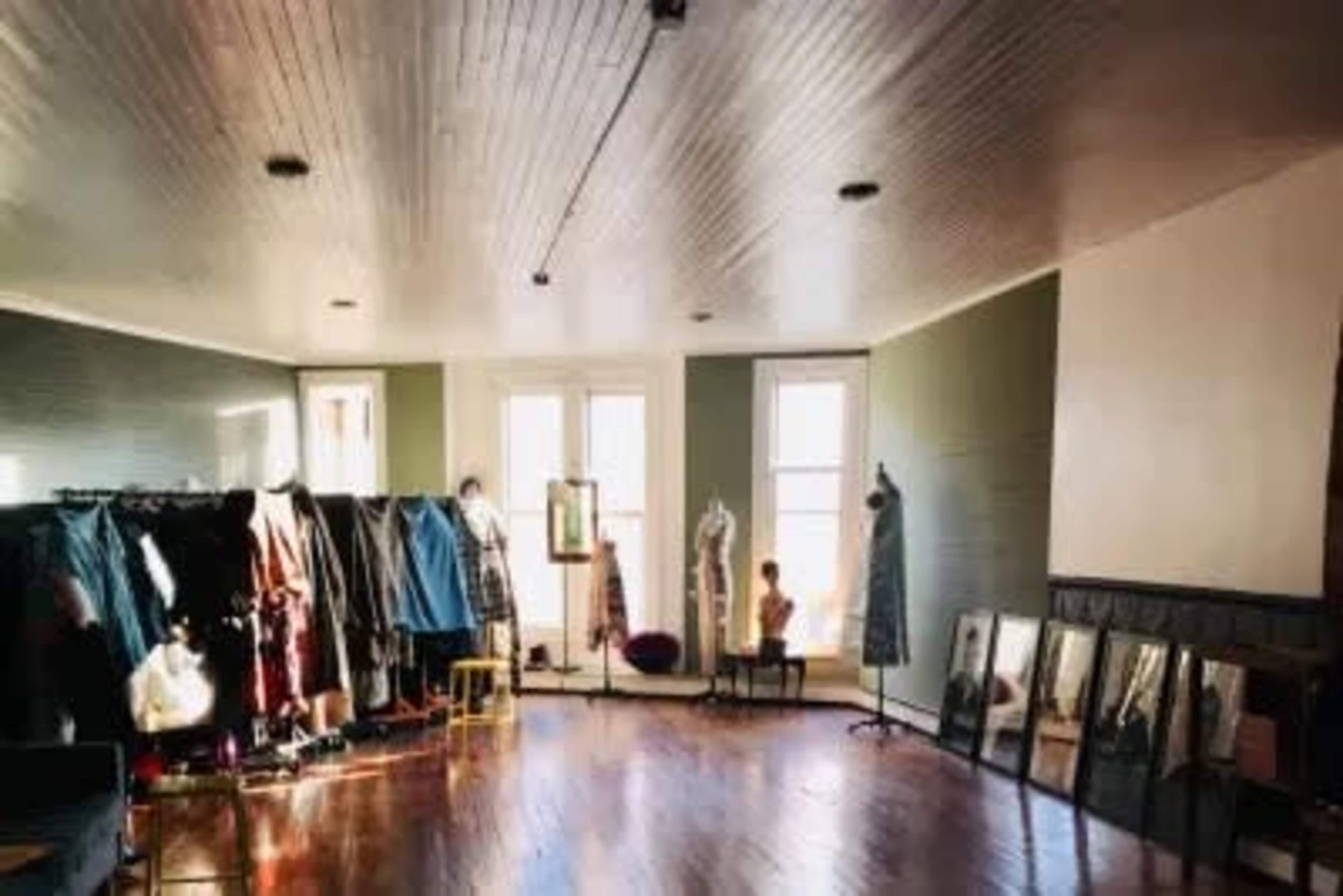 A spacious room features several clothing racks, mirrors, and a mannequin, with large windows allowing natural light to fill the space.