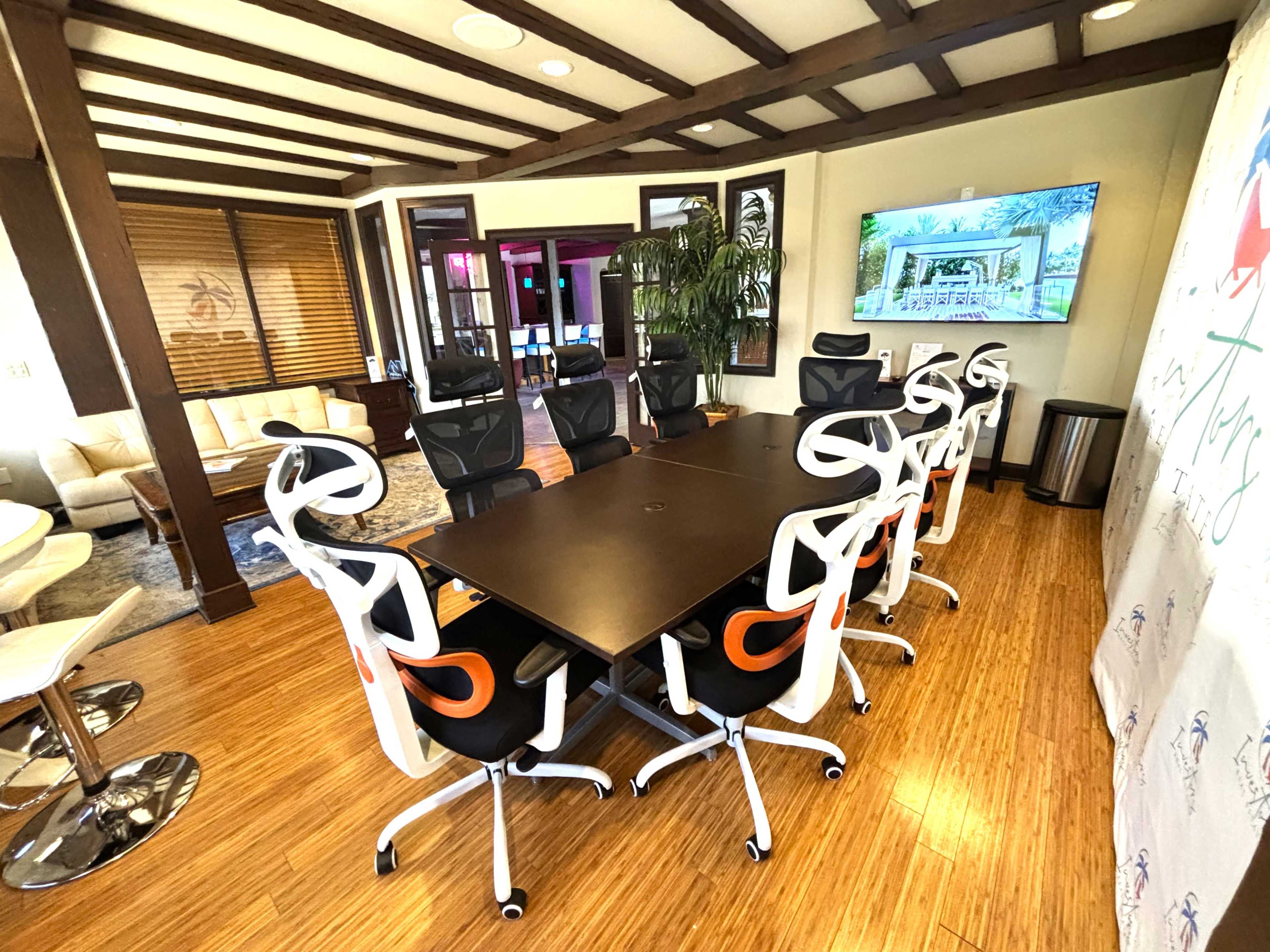 A modern conference room features a long black table surrounded by ergonomic white and orange chairs, with a large screen at one end and large windows providing natural light.