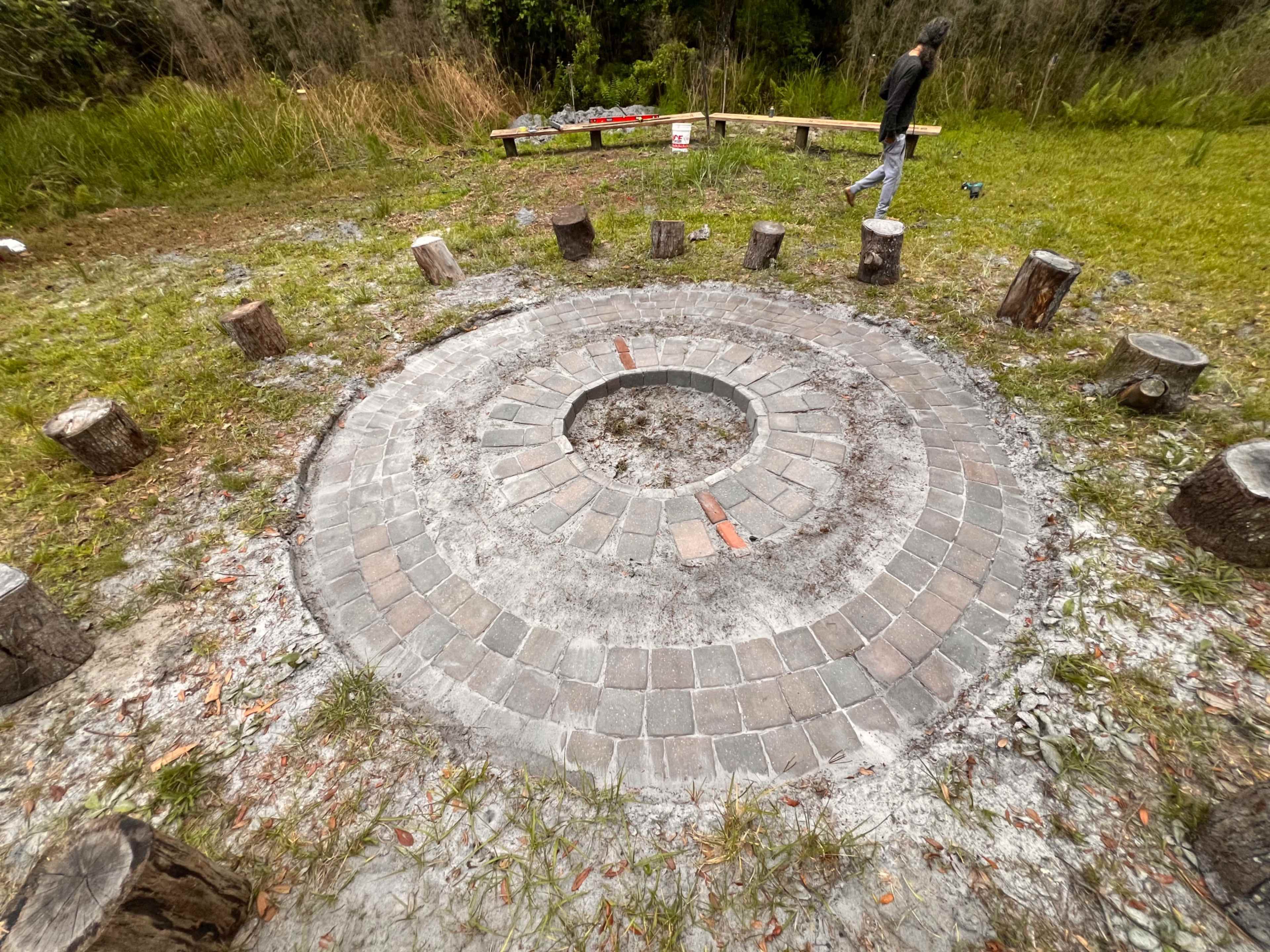 A circular fire pit surrounded by a stone border and wooden logs serves as seating in a grassy clearing.