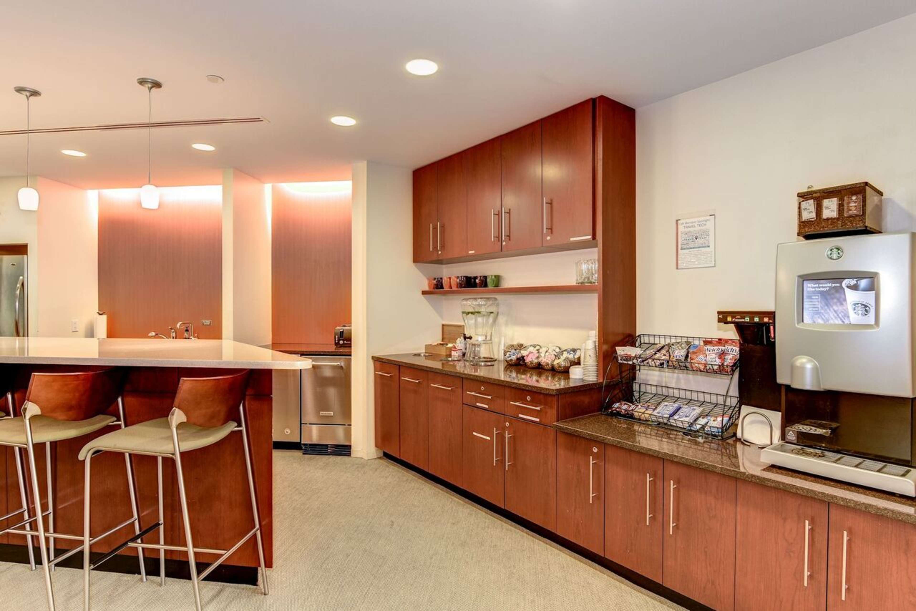 A modern kitchen area featuring wooden cabinets, a countertop with bar stools, a coffee machine, and a snack display.