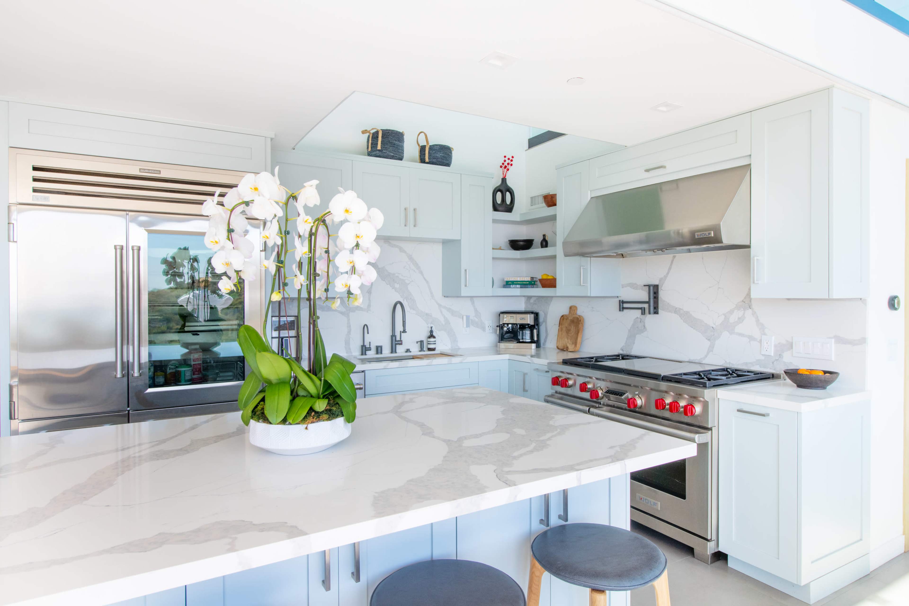 A modern kitchen features light blue cabinetry, stainless steel appliances, and a large marble island with a vase of white orchids.