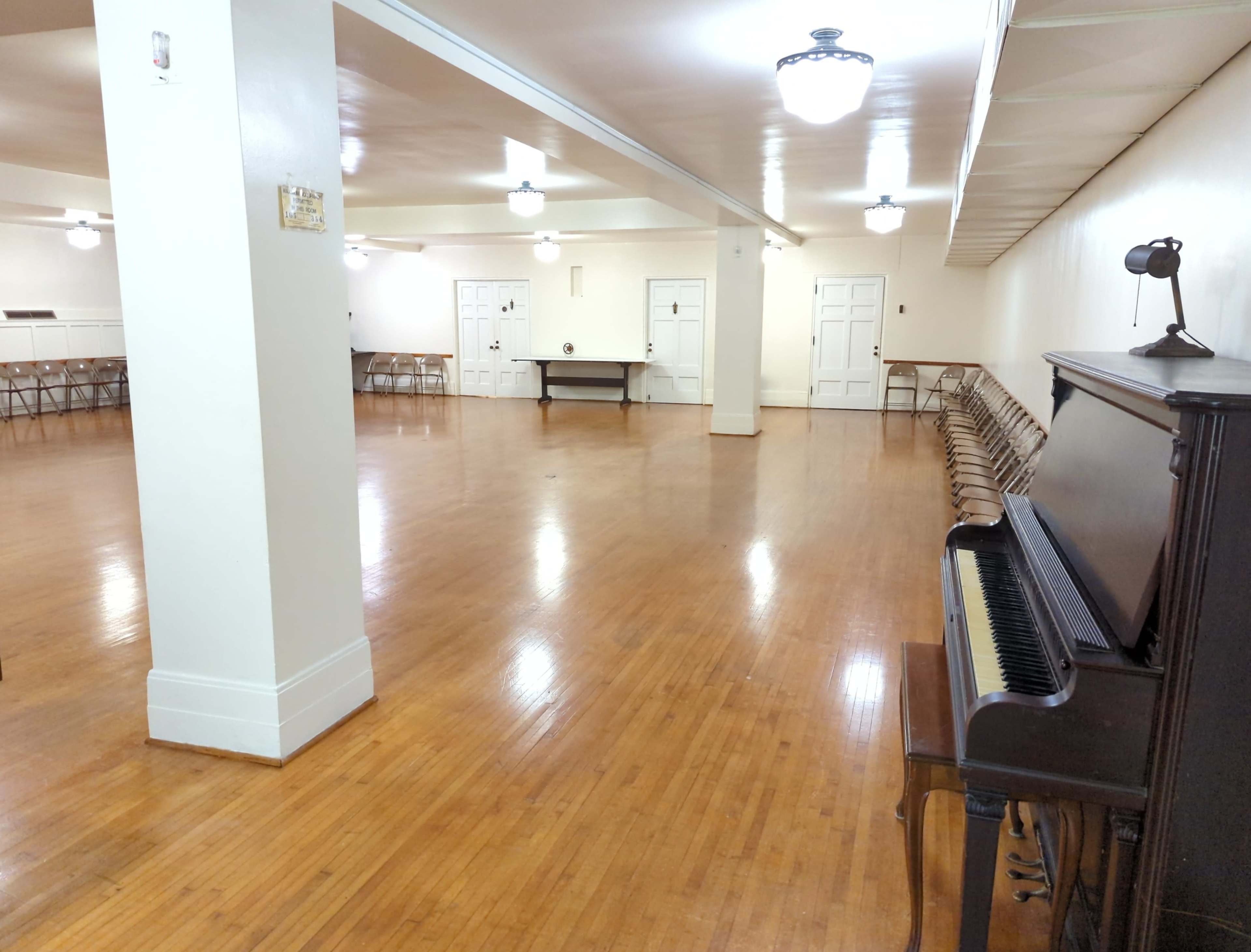 A spacious room with polished wooden floors, several rows of folding chairs, a piano in the corner, and plain white walls.