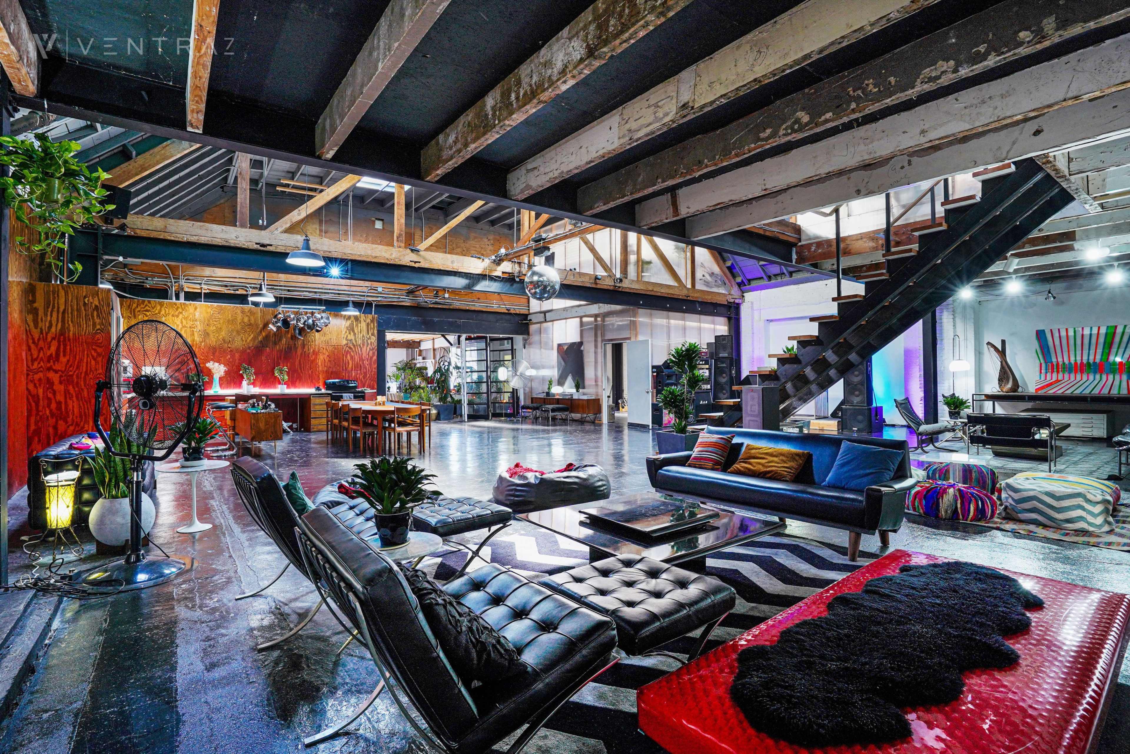 A spacious loft interior with modern furniture, large windows, and exposed wooden beams, featuring a living area, dining space, and a staircase leading to an upper level.