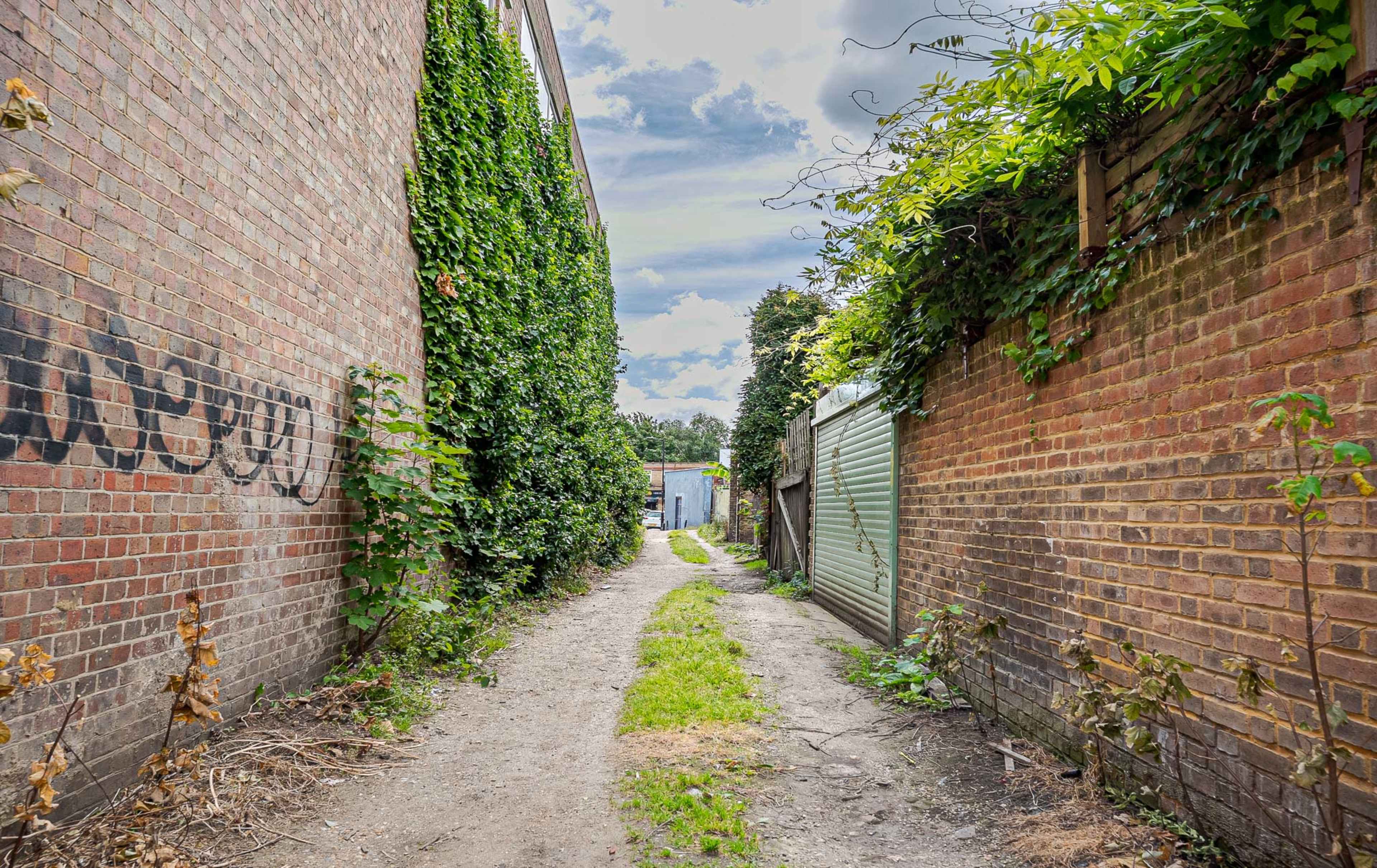A narrow dirt path surrounded by brick walls covered in ivy leads between two buildings.