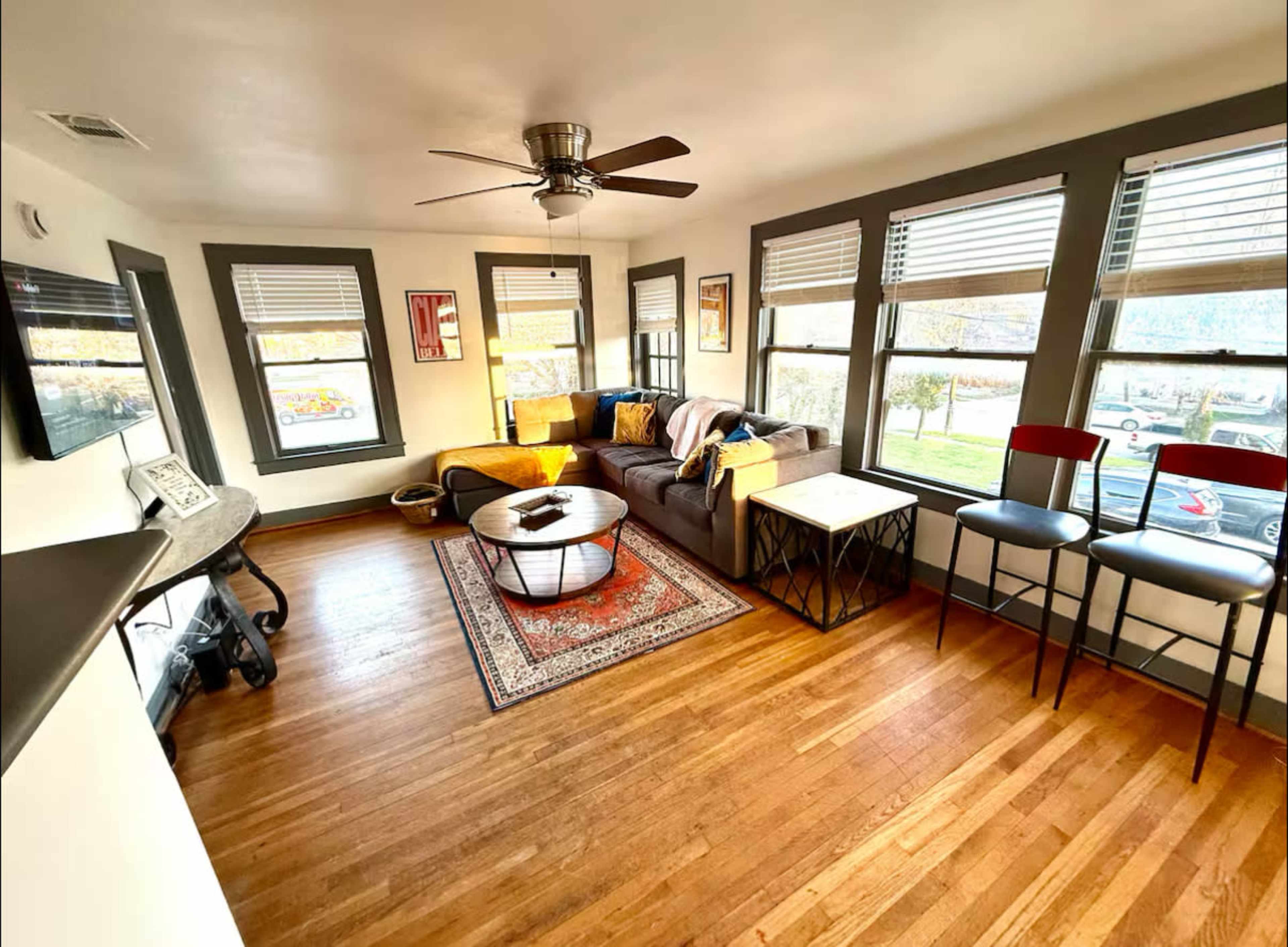 The room features a living area with a gray sofa, a round coffee table, and two bar stools, all illuminated by sunlight streaming through multiple windows.