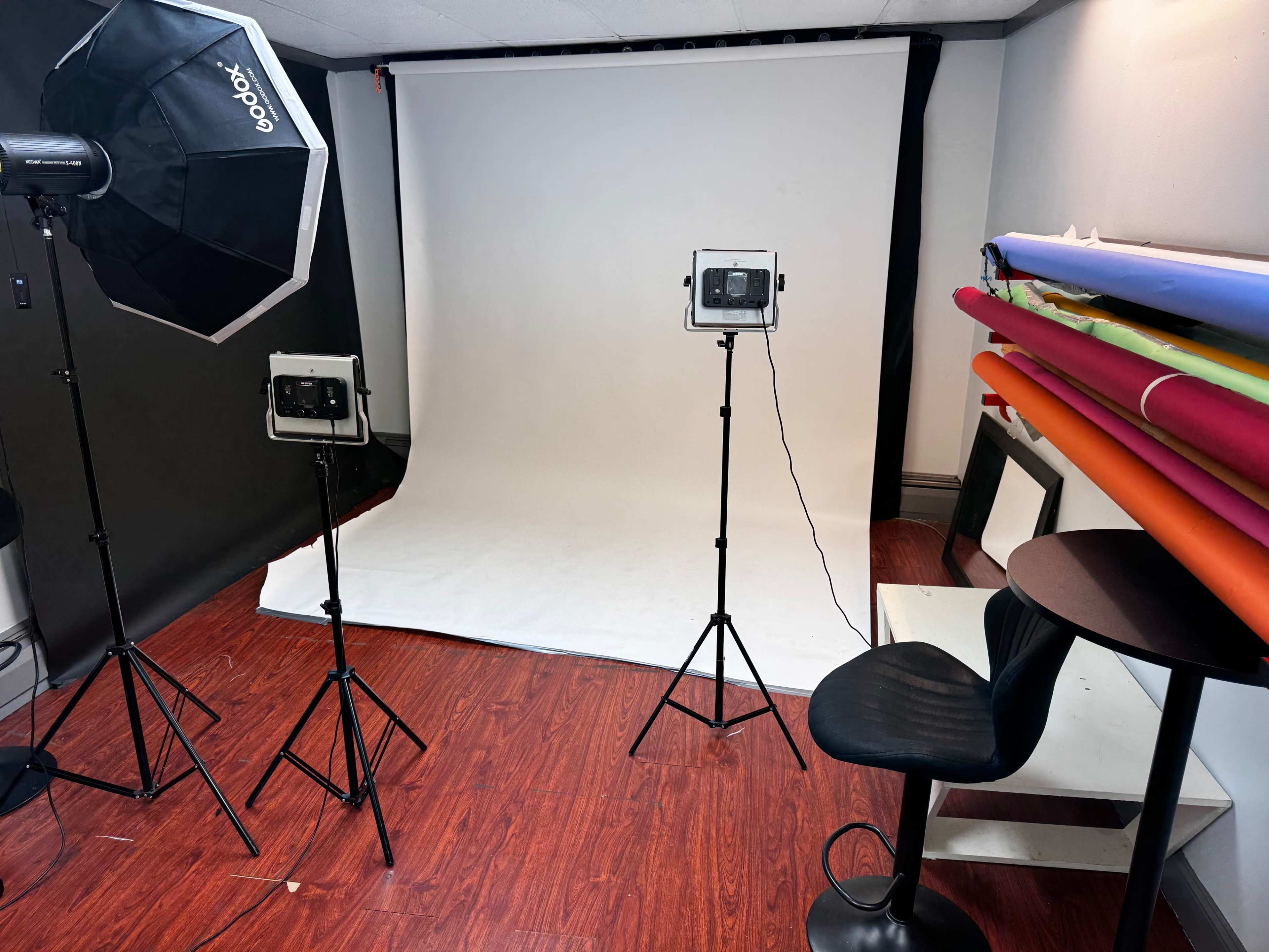 The image shows a photography studio setup with two lighting fixtures, a white backdrop, and a variety of colored backdrops stacked against the wall.