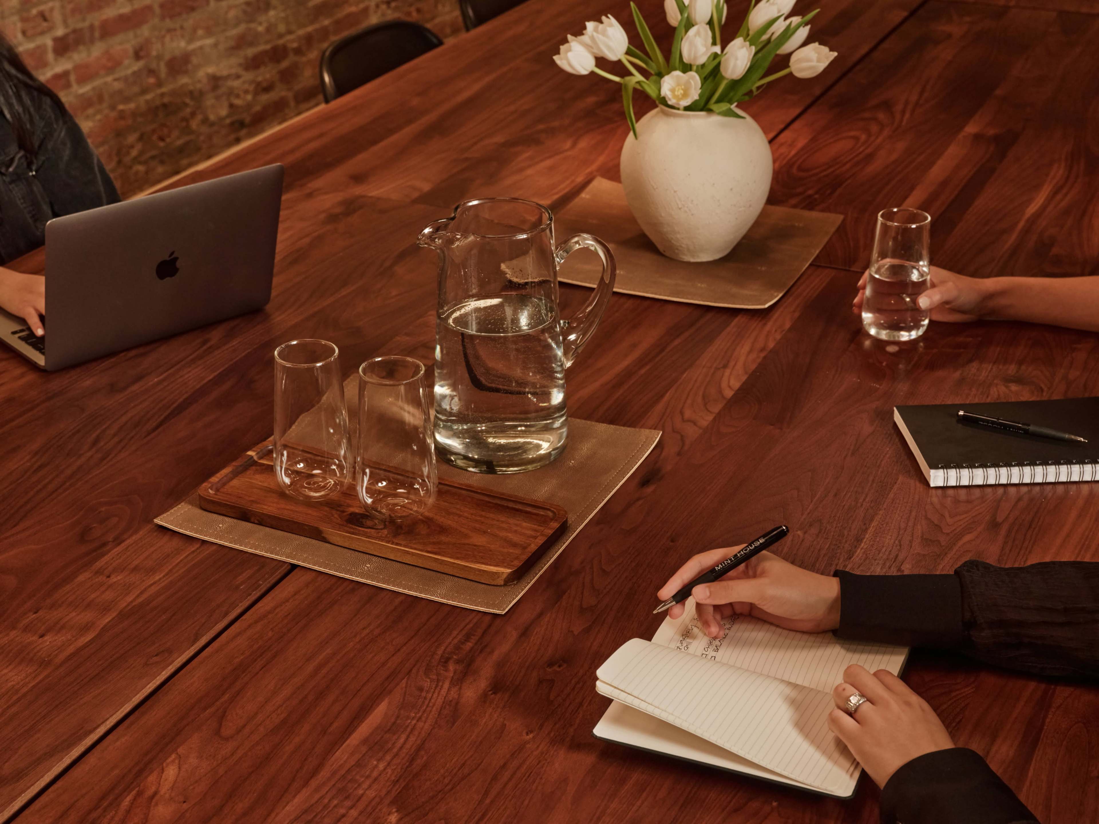 A wooden conference table is set with glasses, a pitcher of water, and a notebook, while two people sit at the sides, one using a laptop and the other writing.
