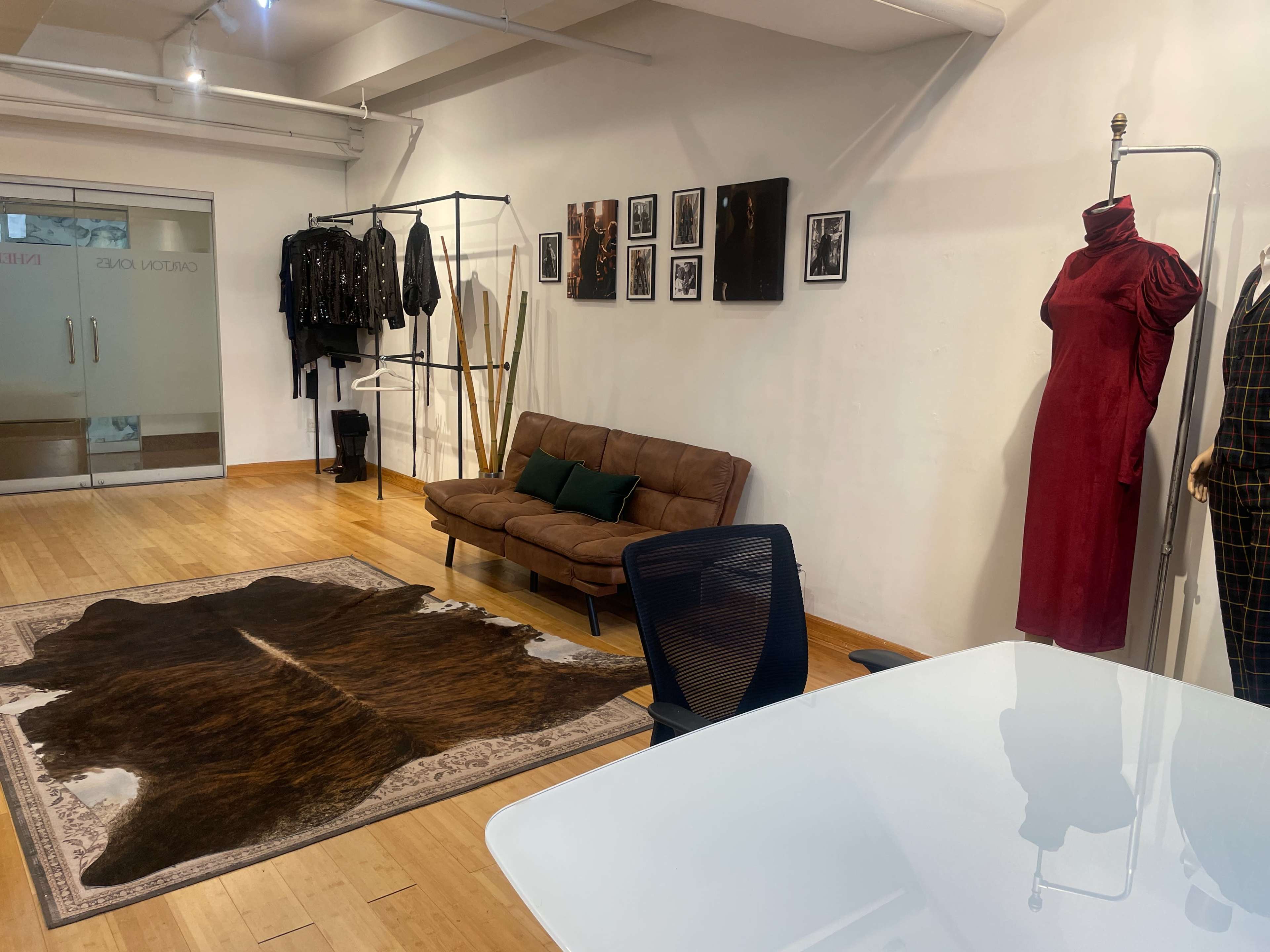 The image shows a modern office space with a brown couch, a desk, a cowhide rug, and clothing displayed on racks and hangers along the walls.