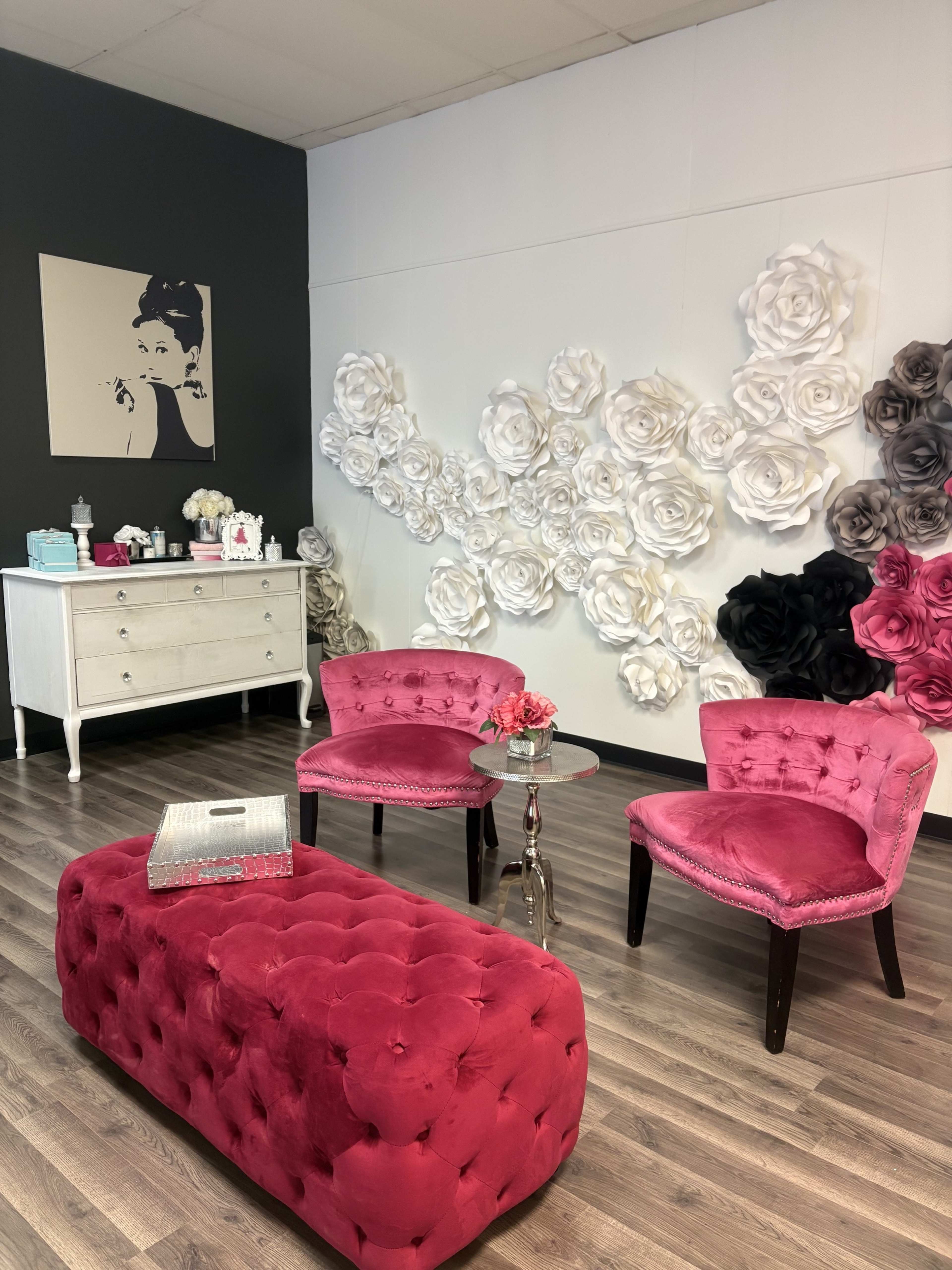 The image shows a stylish interior with two pink velvet chairs, a glass table, and a decorative wall featuring large paper flowers in white, black, and shades of pink.