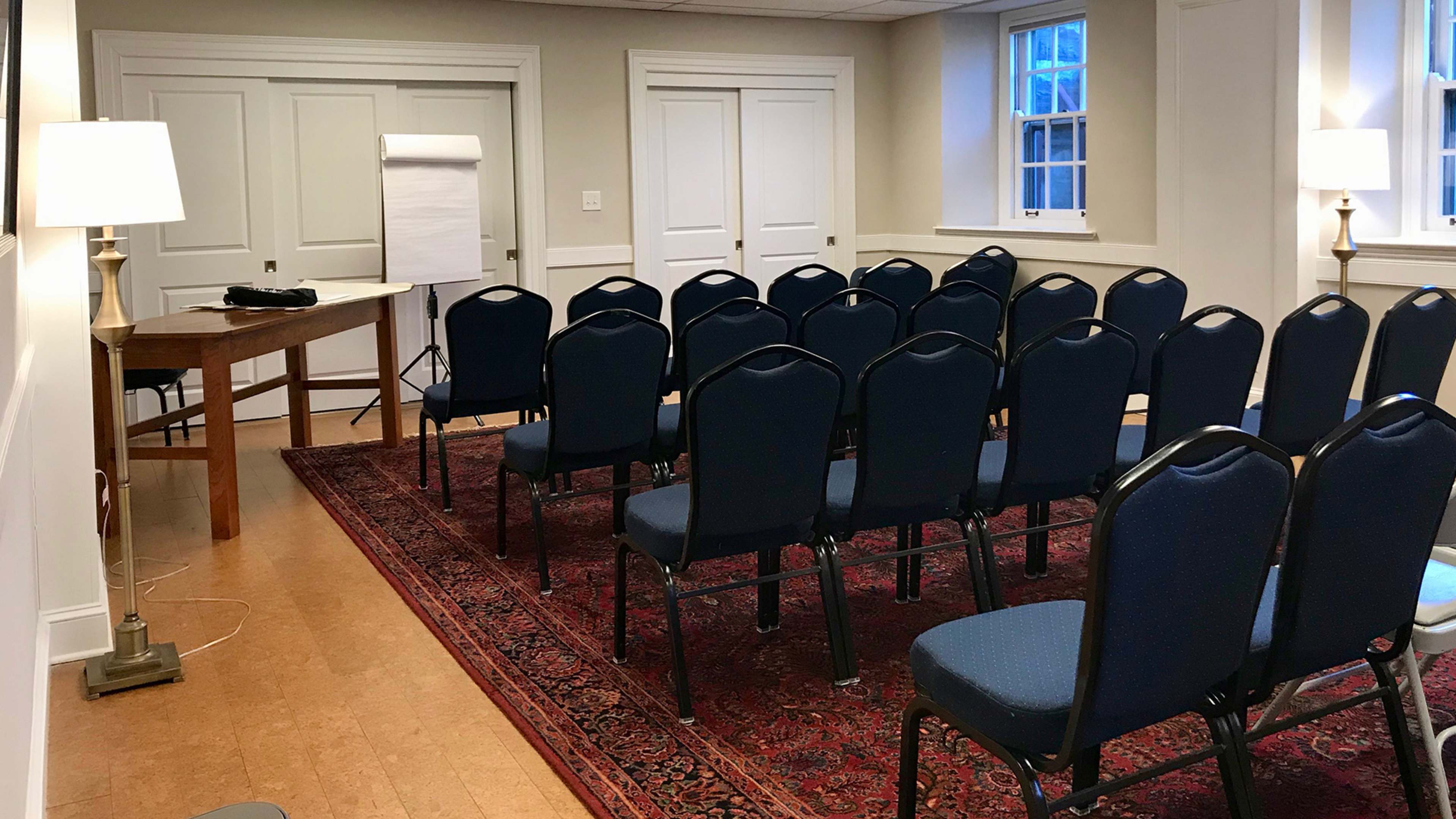 A meeting room features a long wooden table, a whiteboard, and rows of blue chairs arranged on a patterned rug.