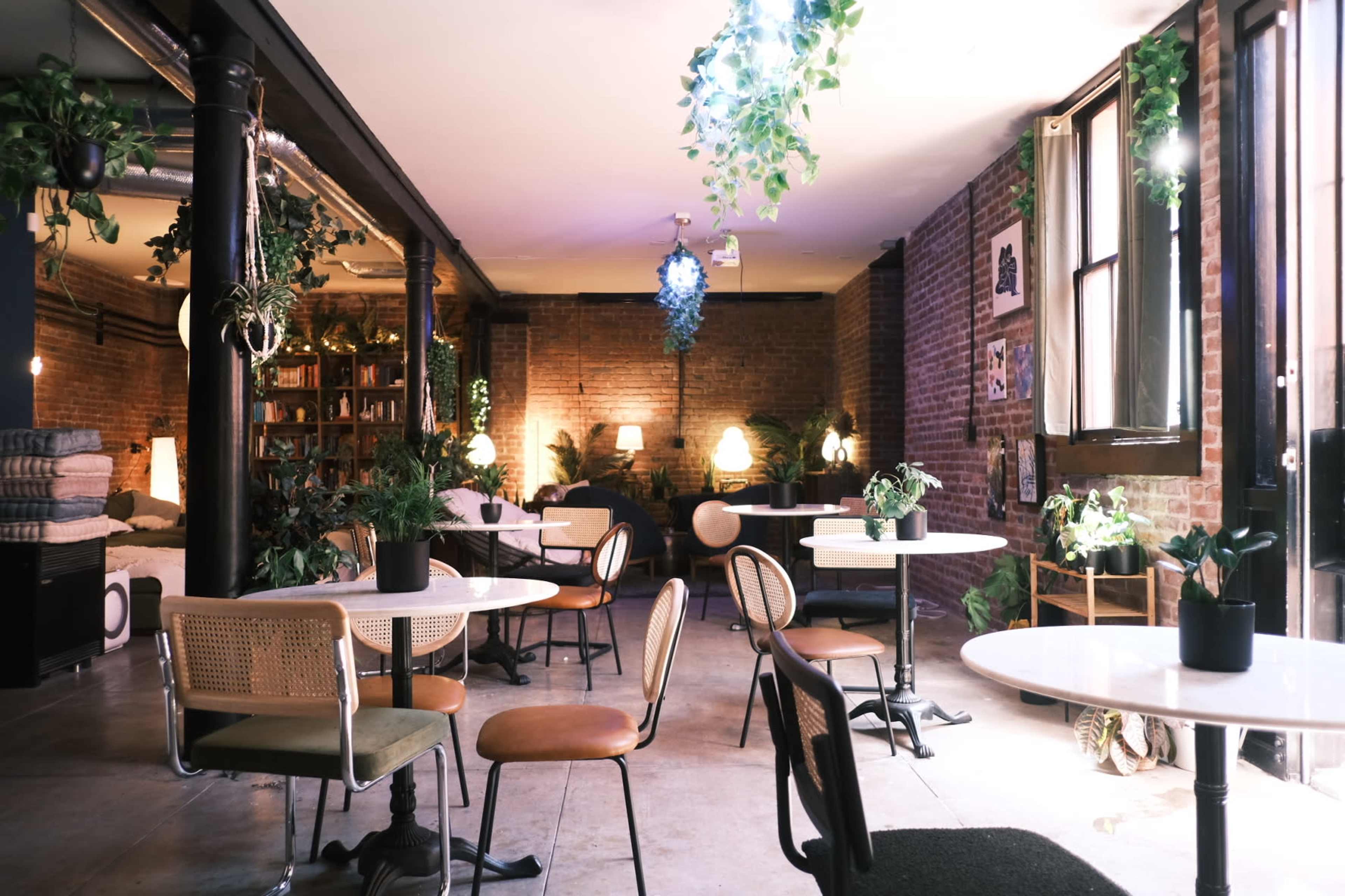 A cozy cafe interior with several tables, chairs, and plants, illuminated by soft lighting against exposed brick walls.