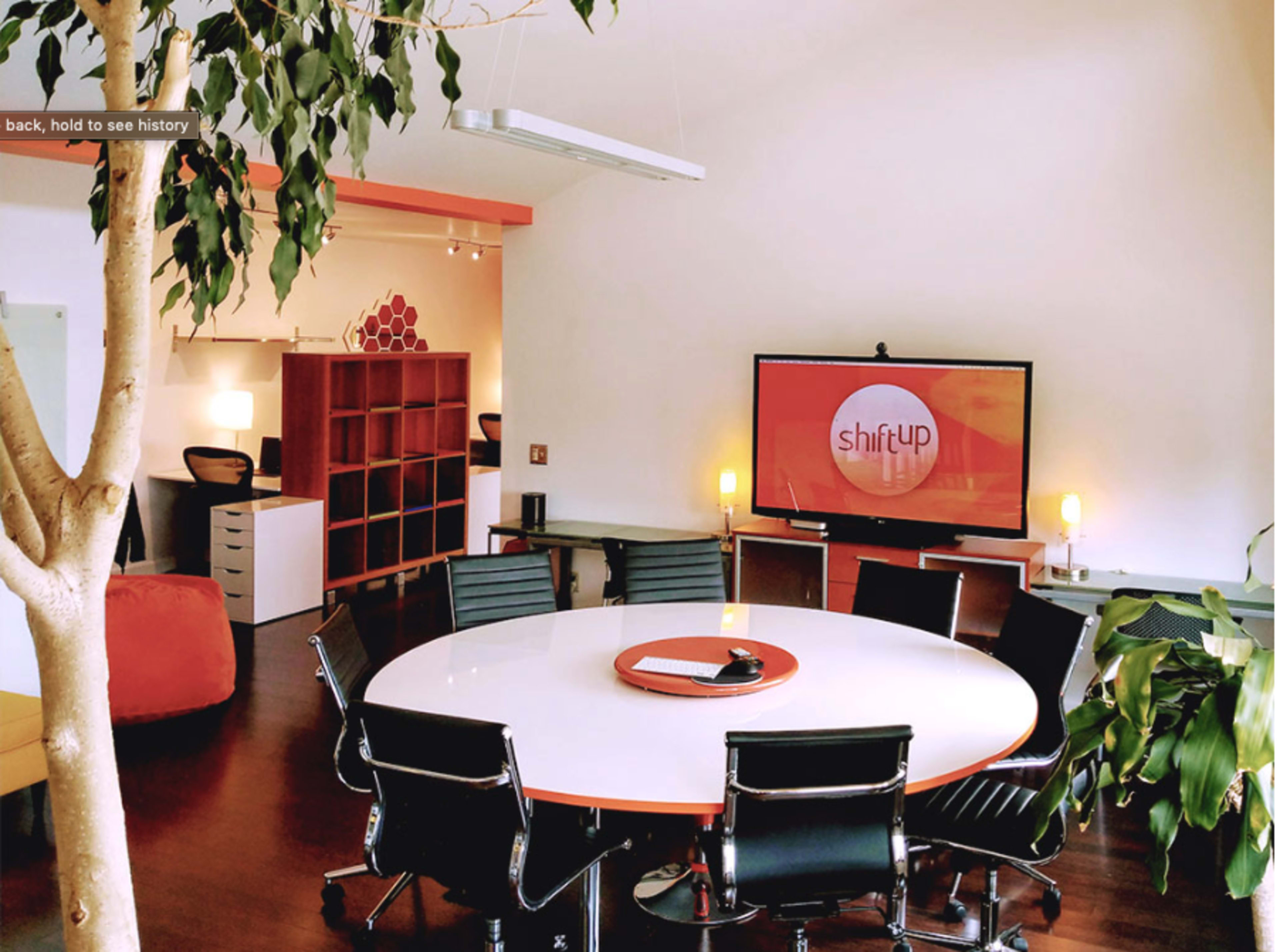 A modern office space features a round white table surrounded by black chairs, with a large screen displaying the logo "shiftUP" on the wall.