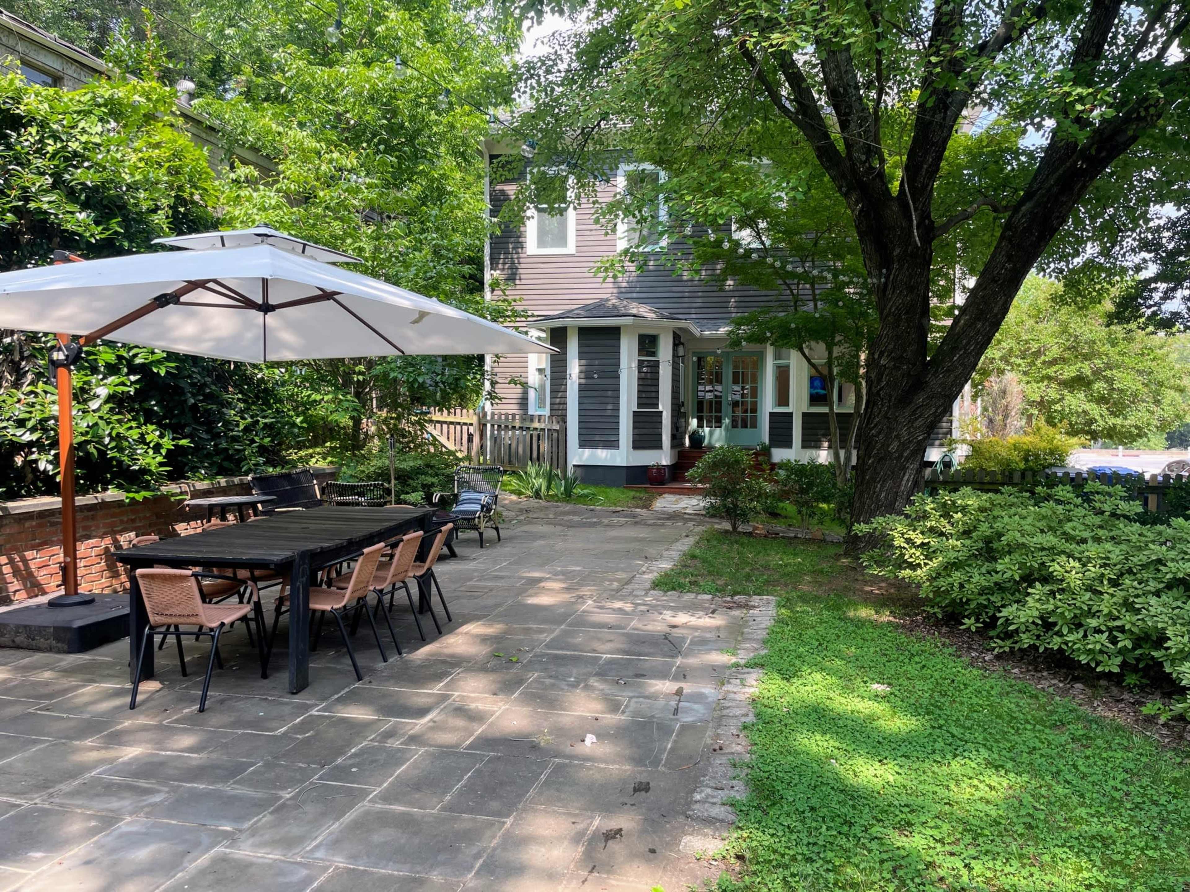 A rectangular patio with a large table, chairs, and an umbrella is set against a house surrounded by greenery and trees.